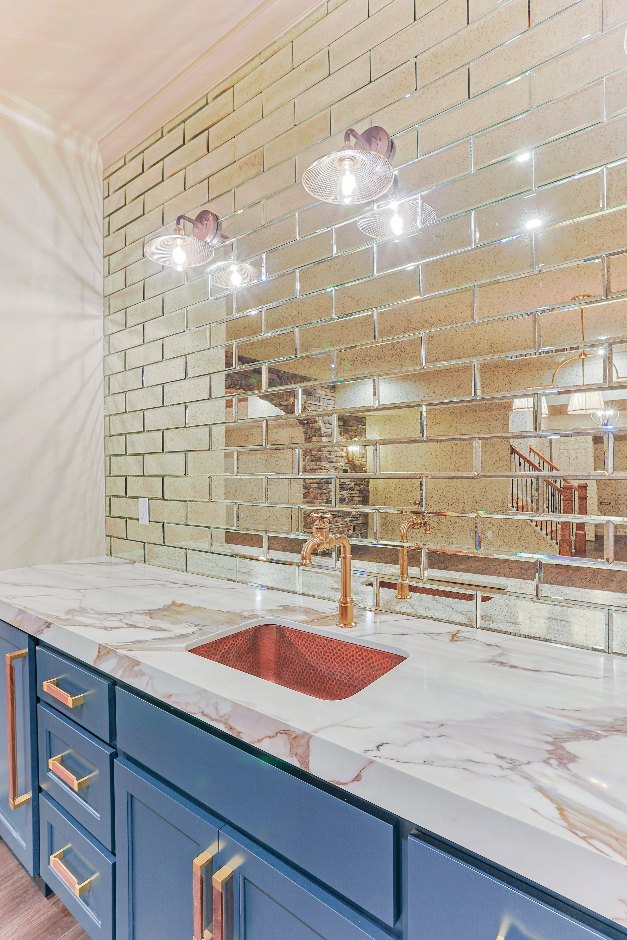 Modern kitchen with a marble countertop, blue cabinets with gold handles, a copper sink, and a mirrored brick wall reflecting the room's interior and staircase.