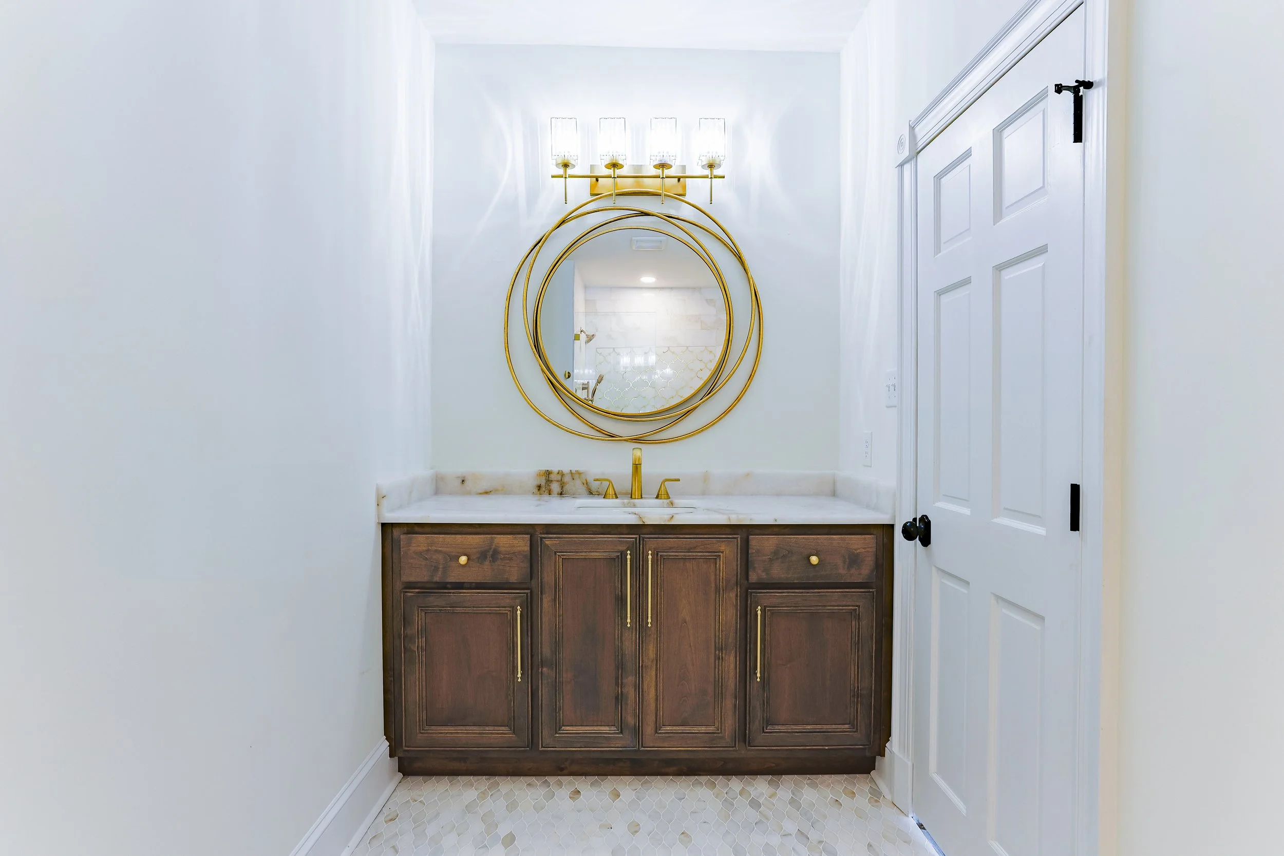 Bathroom vanity with dark wood cabinet, marble countertop, gold faucet, round gold mirror, and a wall-mounted gold light fixture with four glass shades.