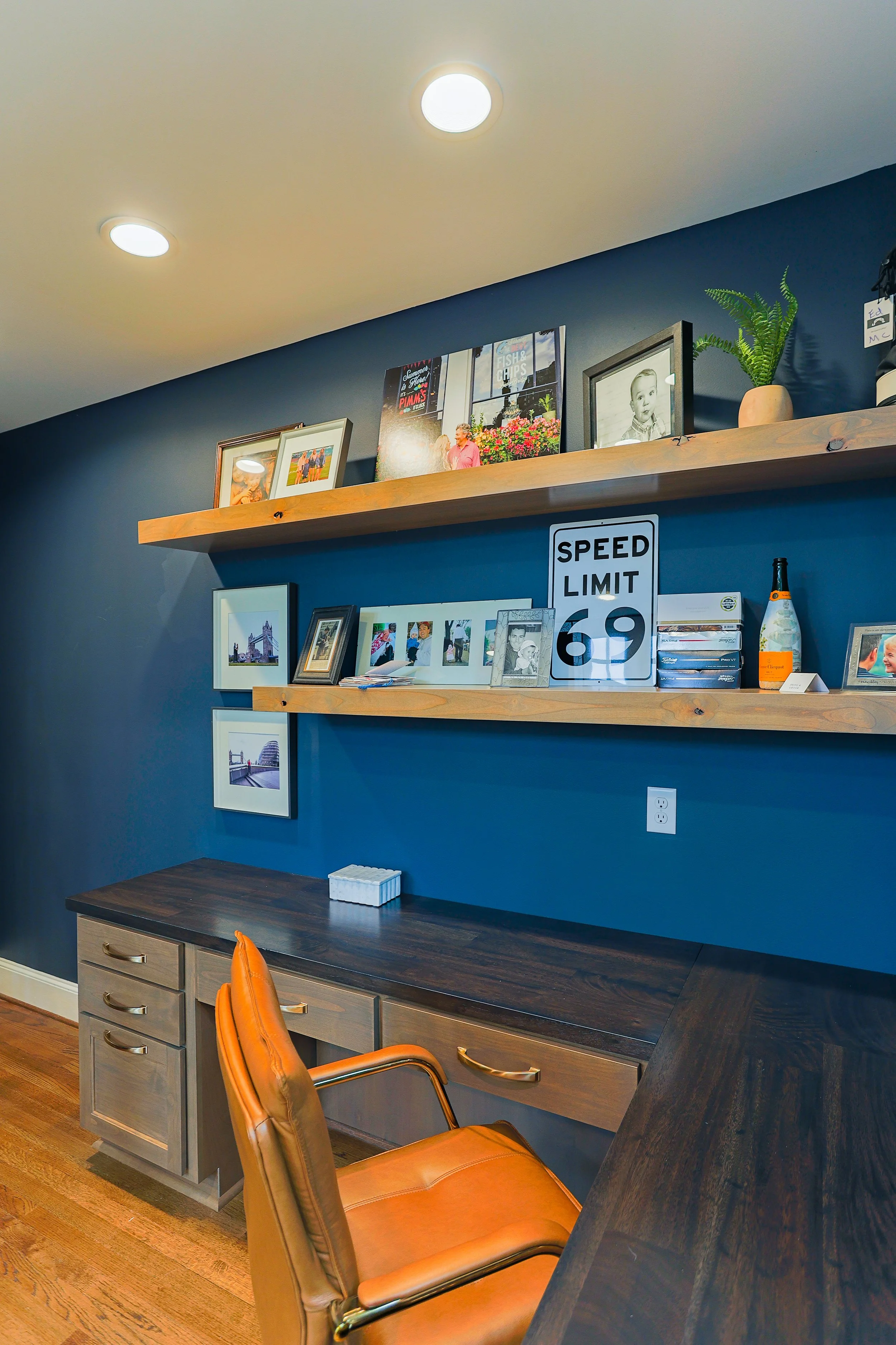 Home office with dark blue wall, wooden shelves with framed photos, a and decorative items, a dark wood desk, and a tan leather office chair.