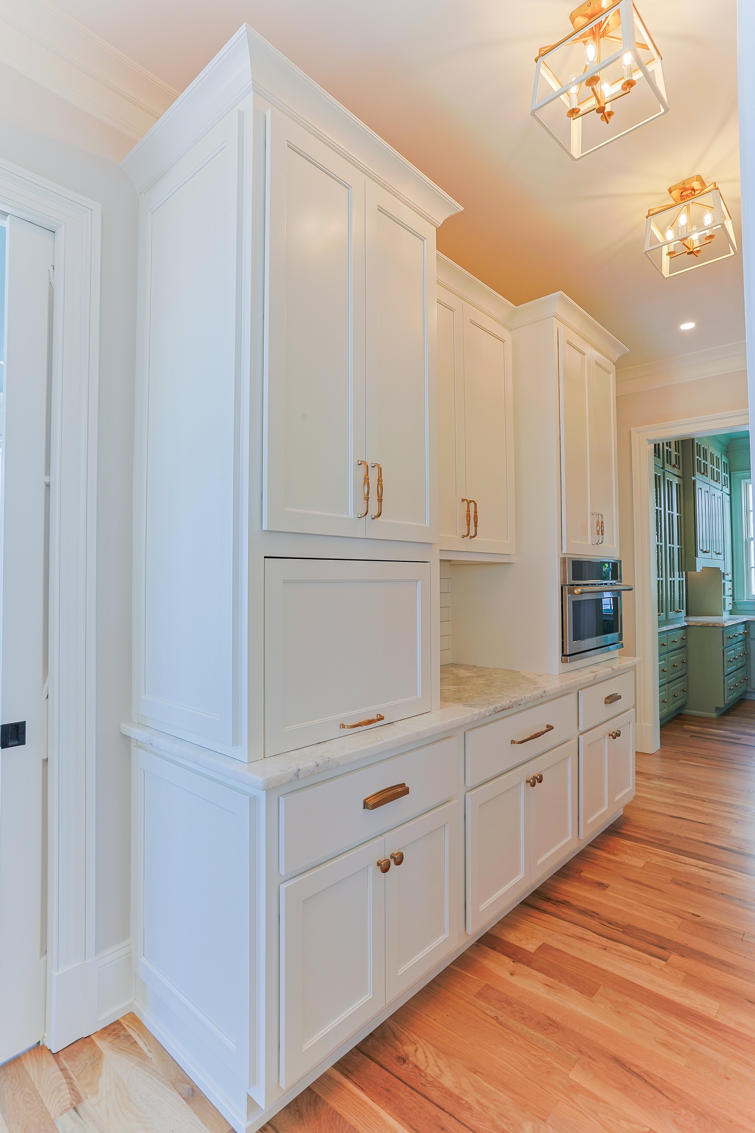 Bright white kitchen cabinetry with gold handles and marble countertops, with hardwood floors and modern hanging light fixtures.