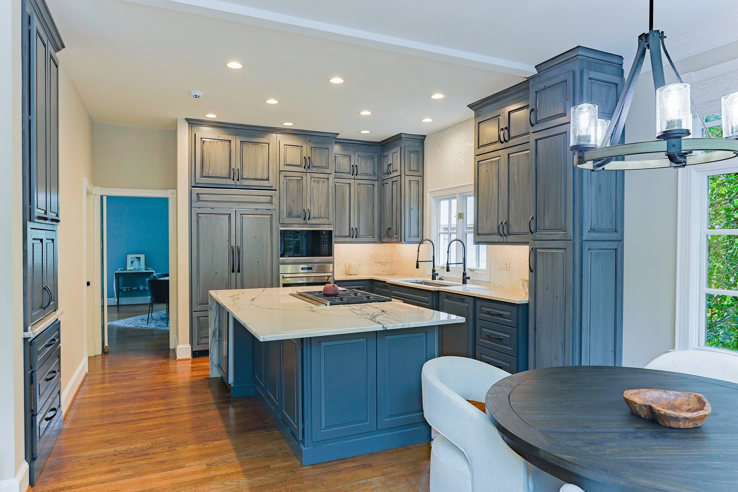 Modern kitchen with blue cabinetry, marble island, hardwood floors, and a dining area with dark wood table and white chairs.