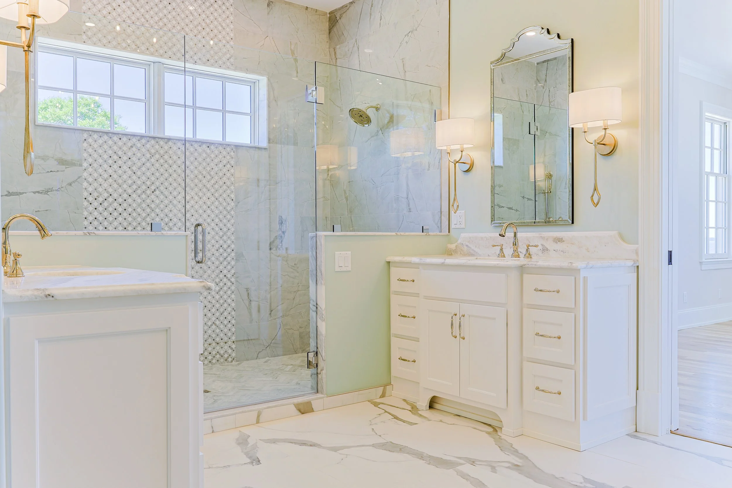 Bright bathroom with white cabinets, marble countertops, a large mirror, wall-mounted lights, a glass-enclosed shower with patterned tile, and a window with natural light.