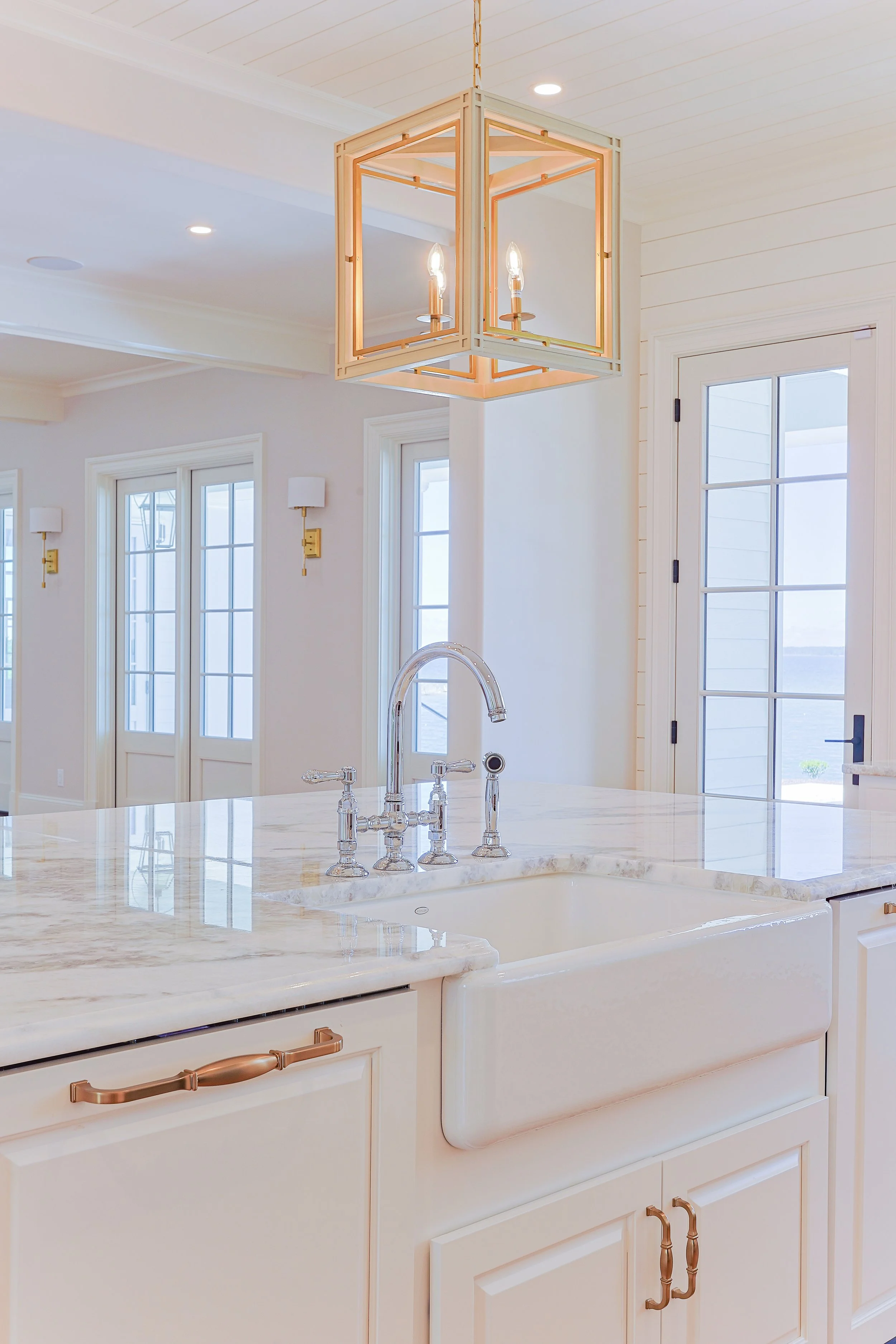 Bright kitchen with white cabinets, a farmhouse sink, marble countertop, and a geometric pendant light fixture hanging from a white ceiling. Multiple windows and a glass door provide natural light.