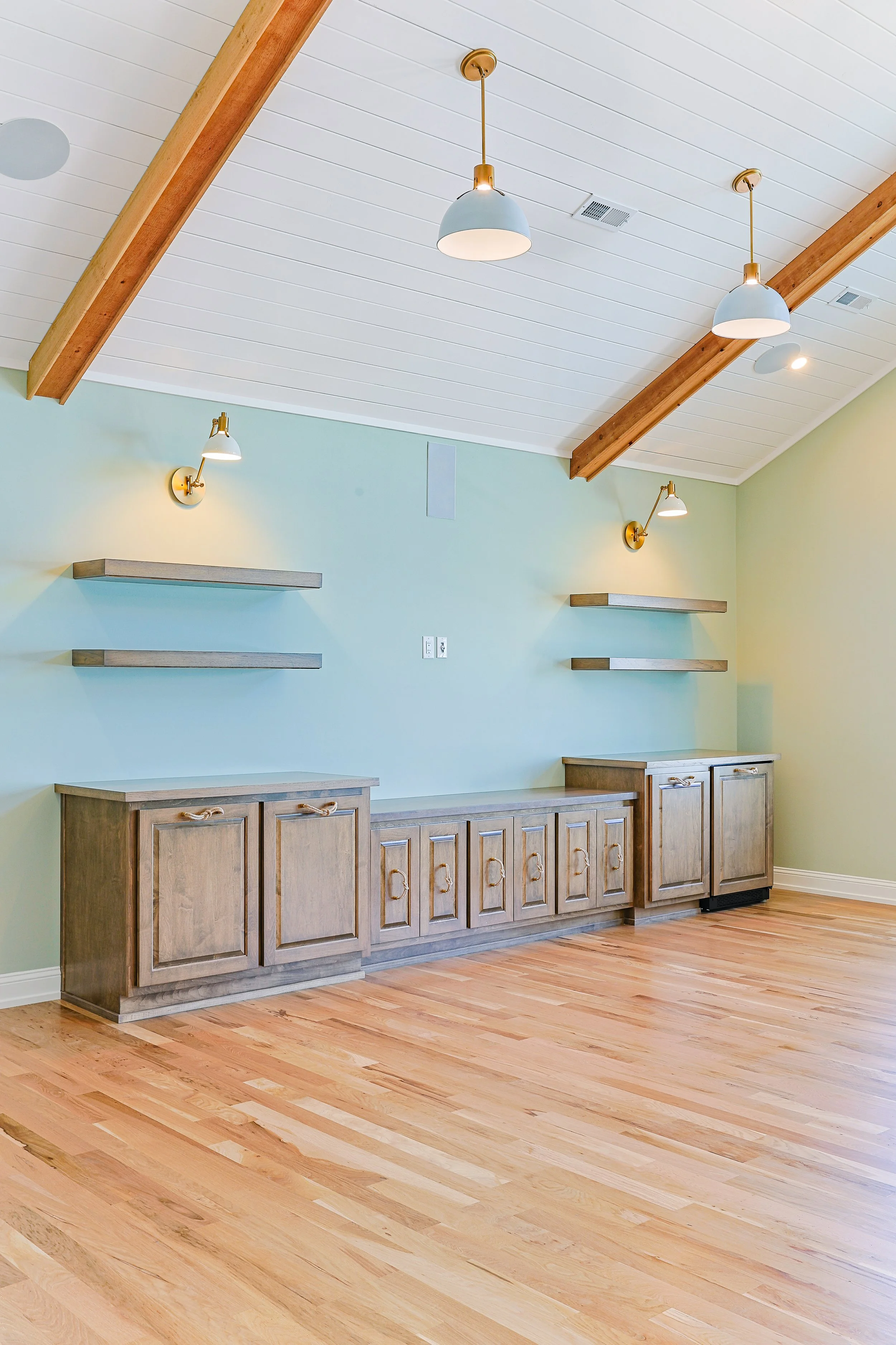 Empty room with vaulted ceiling, light green walls, wooden cabinets, floating shelves, and hardwood floors.