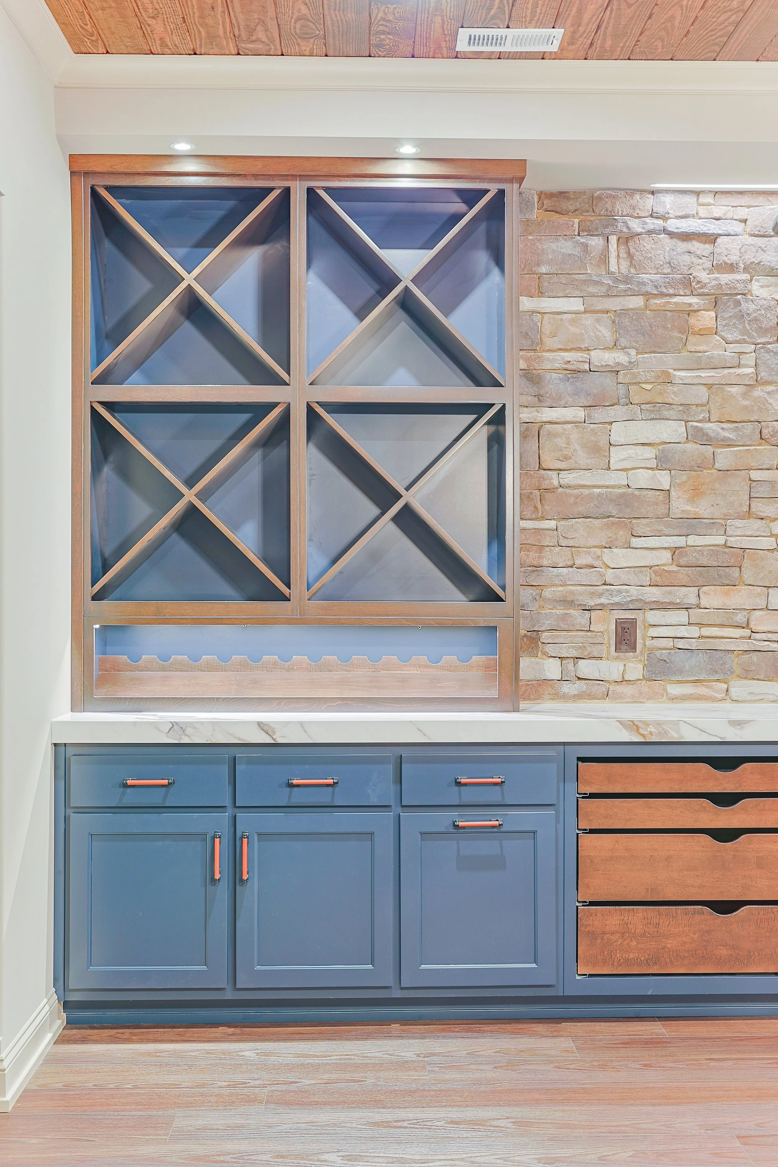 A kitchen cabinet with blue doors, a marble countertop, and wooden drawers, set against a brick wall and wooden ceiling.