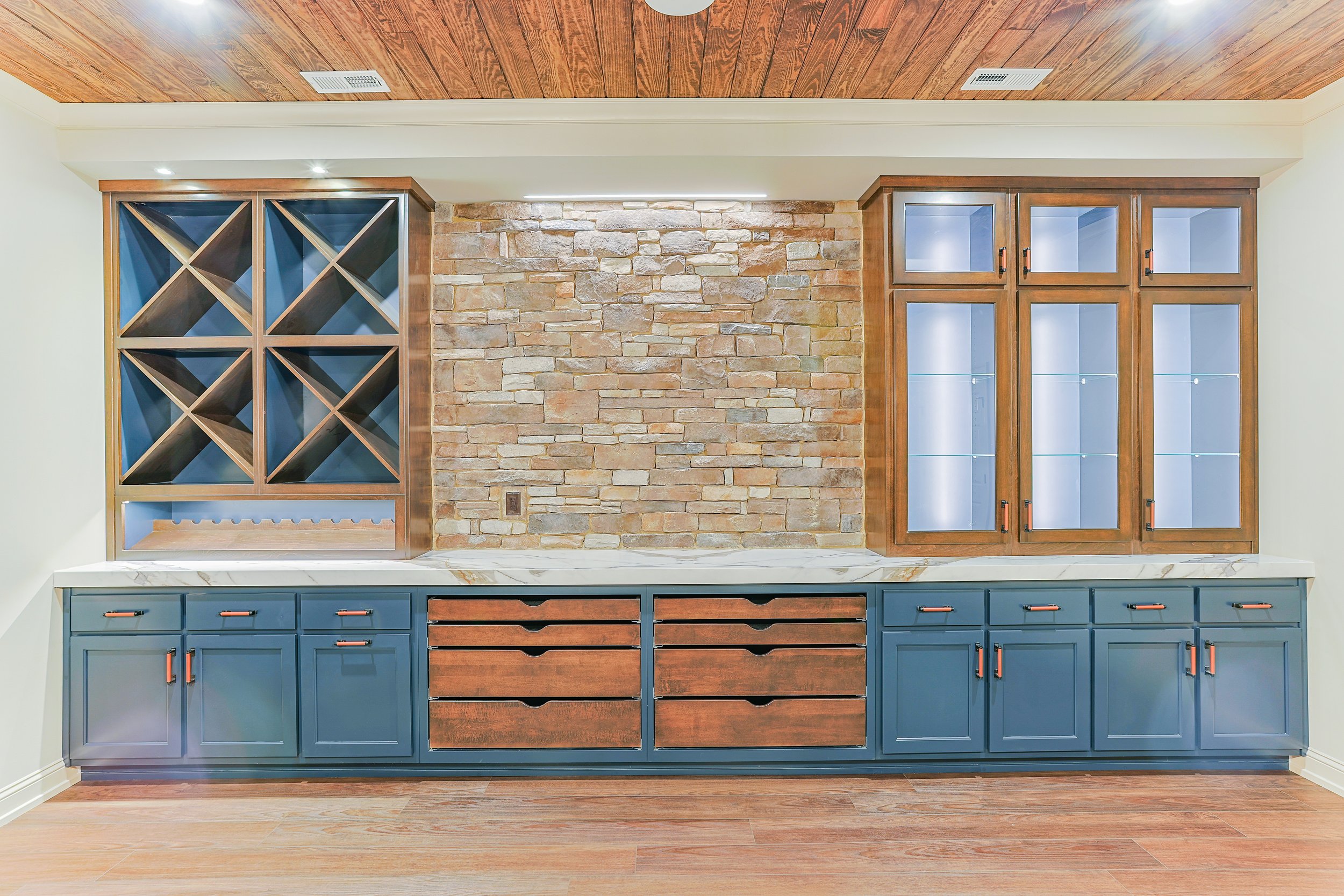 Kitchen bar area with blue cabinets, wooden wine rack, and glass-front display cabinets, with a stone accent wall and wooden ceiling.