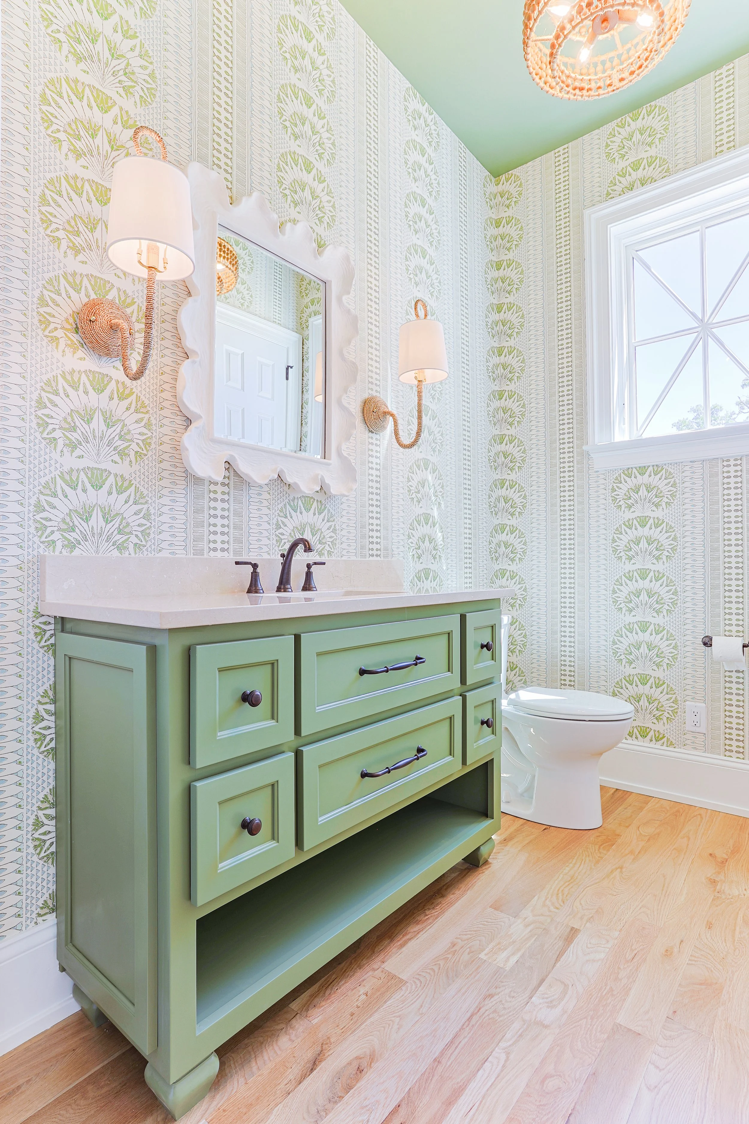 A bathroom with a green vanity, wall-mounted lamps, a mirror, a window, and wallpaper with green and beige patterns.