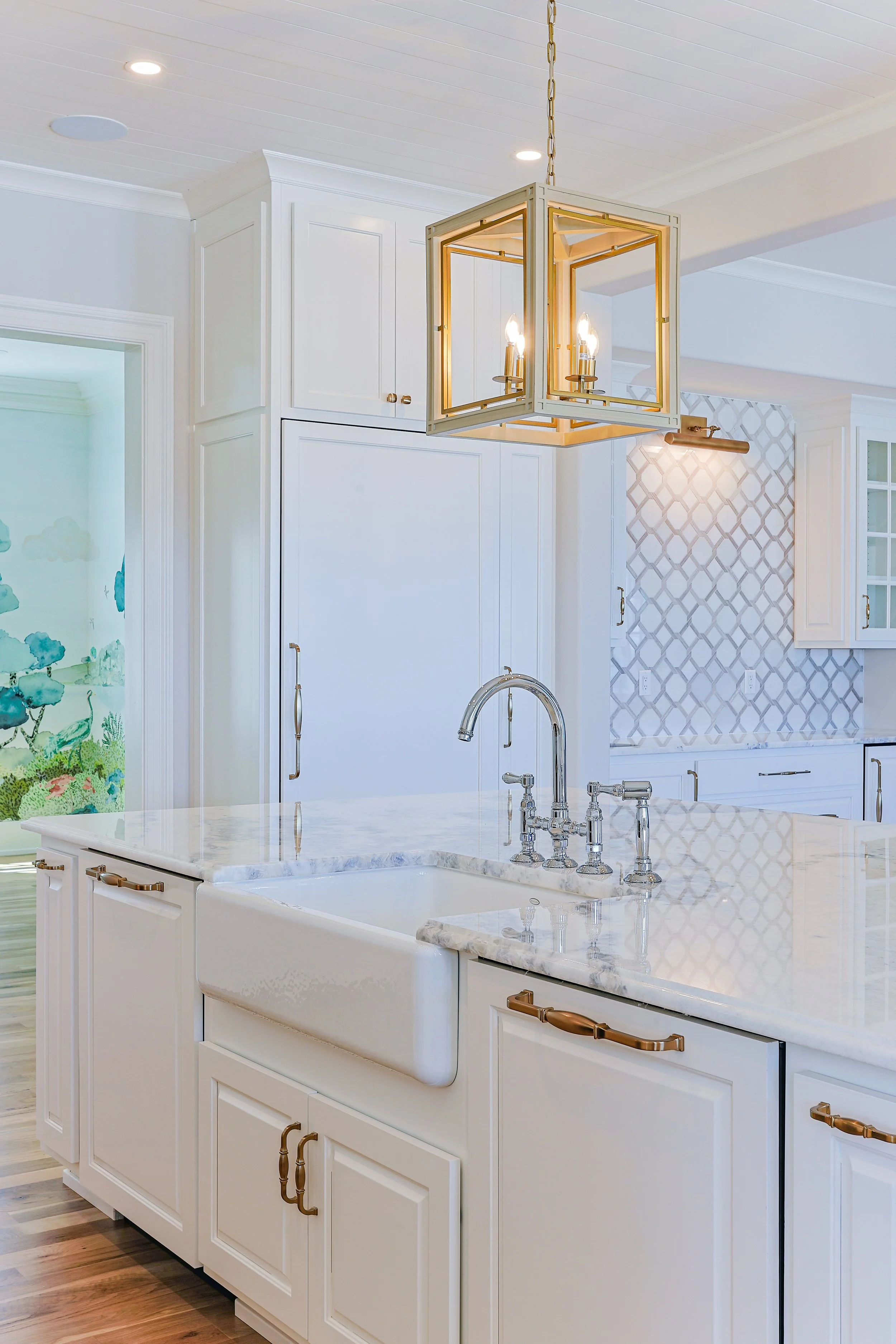White kitchen with marble countertop, gold handles, farmhouse sink, gold hanging light fixture, and patterned tile backsplash.