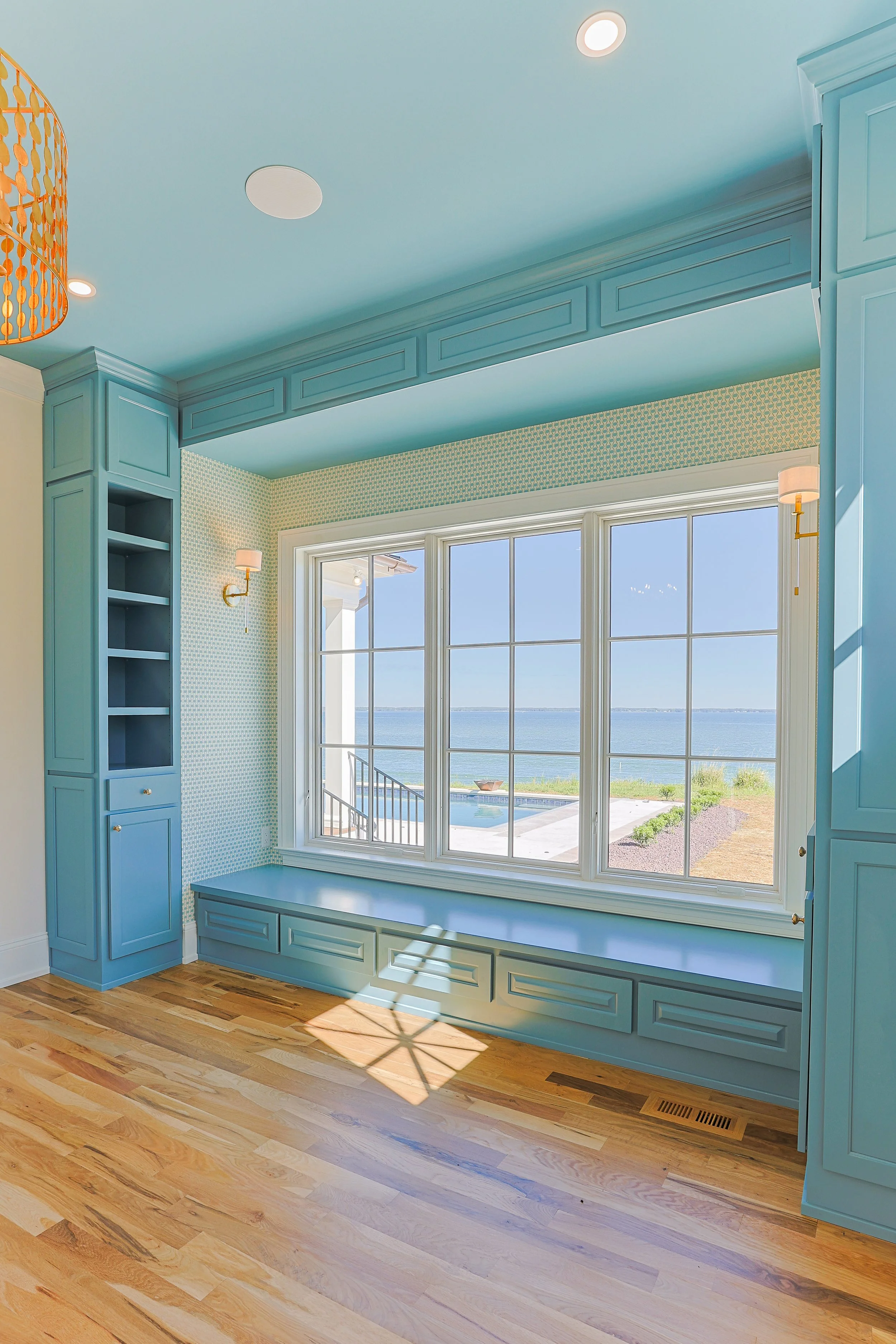 Room with large window overlooking water, built-in blue window seat and cabinetry, hardwood floors, and light blue ceiling.