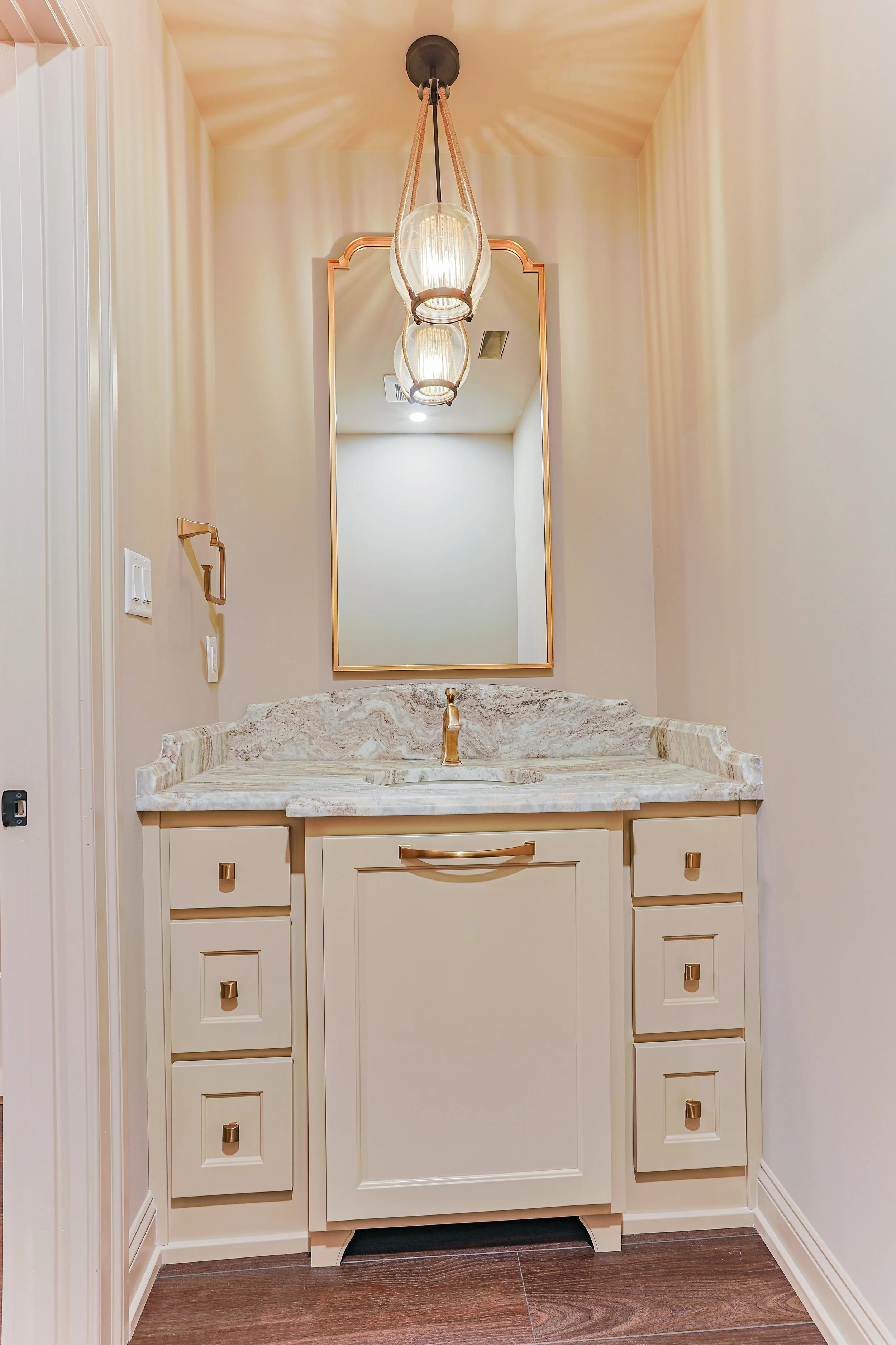 A bathroom vanity with a beige cabinet, a marble countertop, a gold faucet, and a large mirror above it. A pendant light hangs from the ceiling, casting decorative shadows on the ceiling. The walls are painted light cream, and the floor is dark wood.