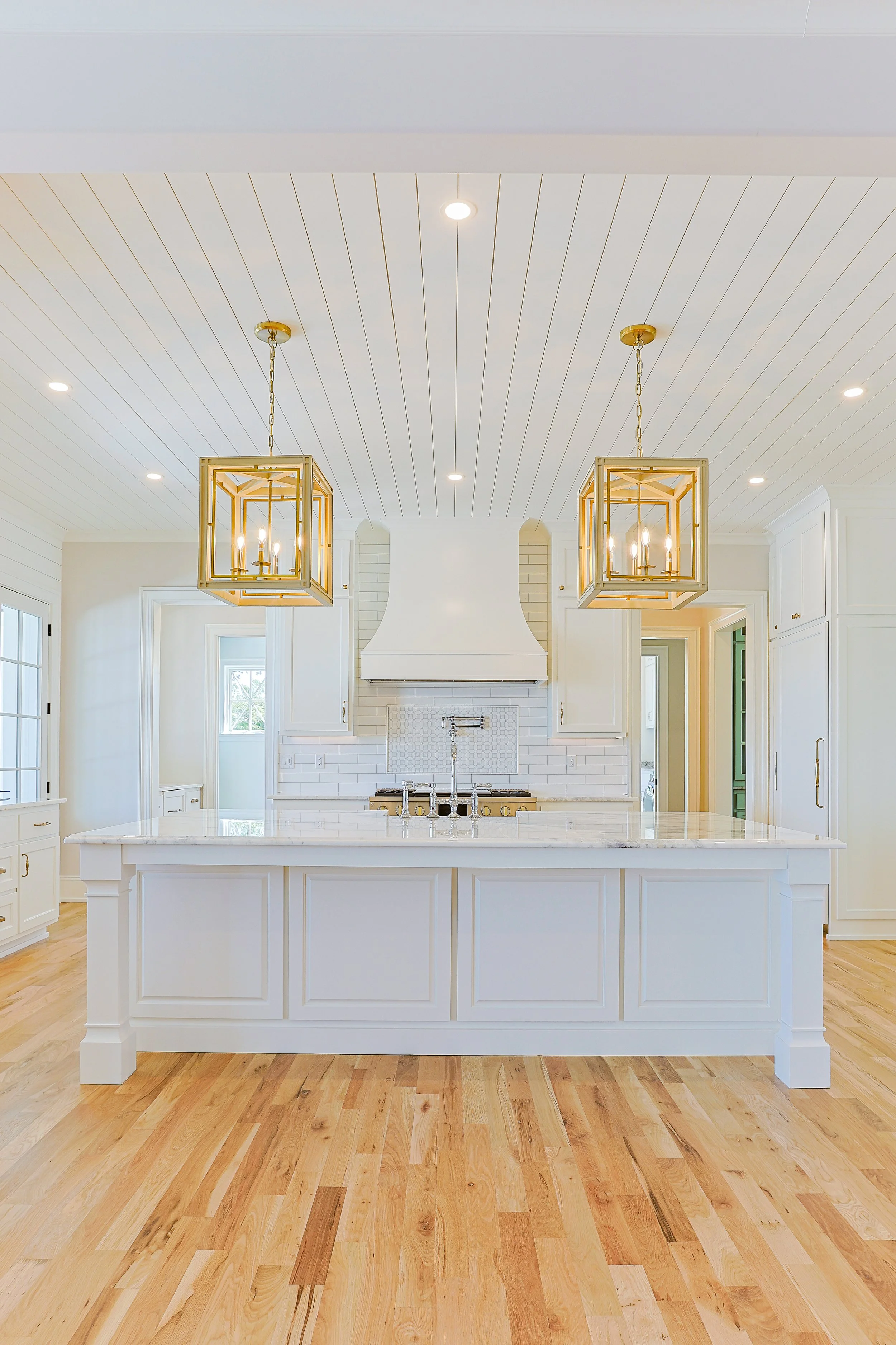 A bright, modern white kitchen with a large island, hanging gold light fixtures, white cabinetry, and hardwood floors.