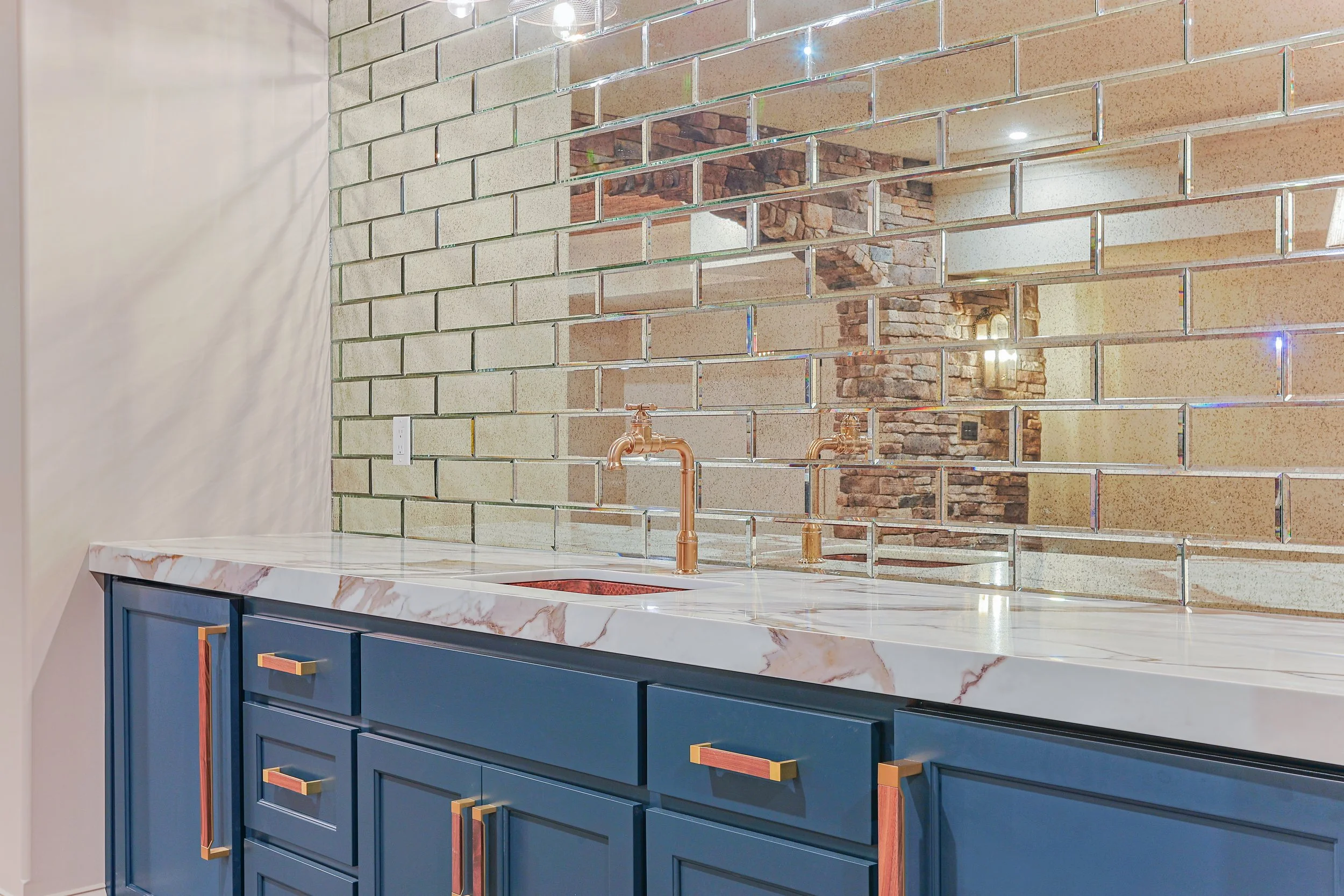 A modern kitchen with a white marble countertop, blue cabinets with copper handles, a brick and mirrored wall, and a copper faucet.