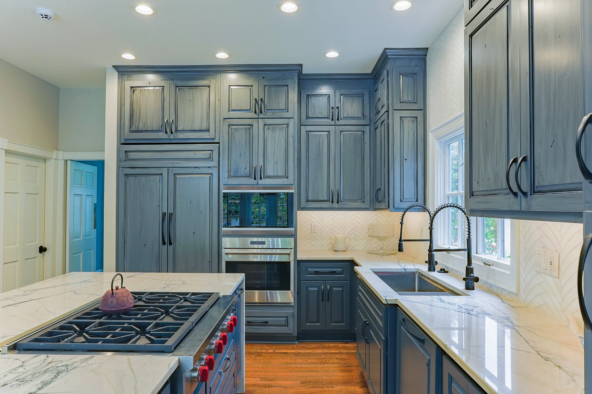 Modern kitchen with blue wooden cabinets, marble countertops, stainless steel appliances, a double sink, a window above the sink, and hardwood floors.
