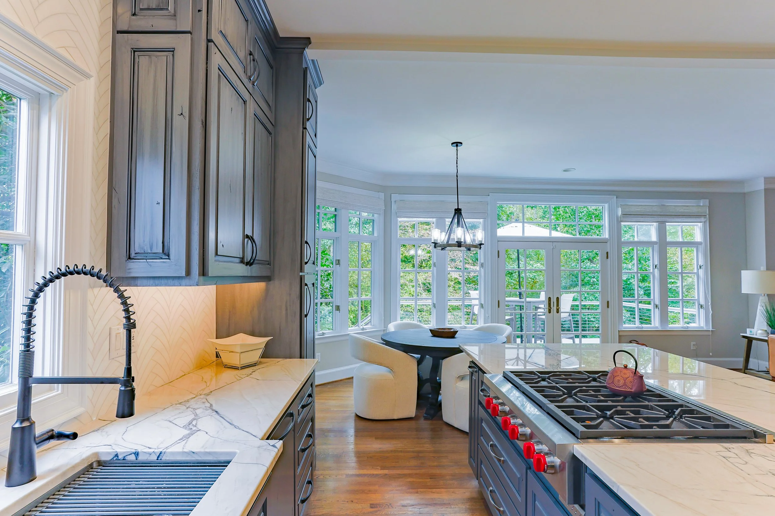 Bright kitchen with dark cabinets, marble countertops, and large windows overlooking greenery. A round dining table with white chairs is in front of a sliding glass door, and a modern chandelier hangs above.