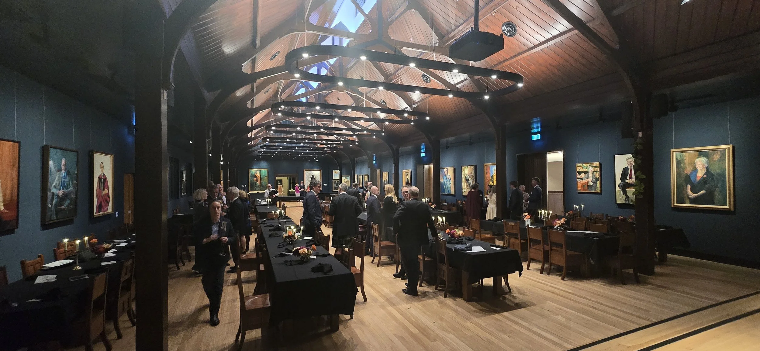 People gathered at Trinity College Hall with painted portraits hanging on dark blue walls, a wooden vaulted ceiling, and modern circular light fixtures.