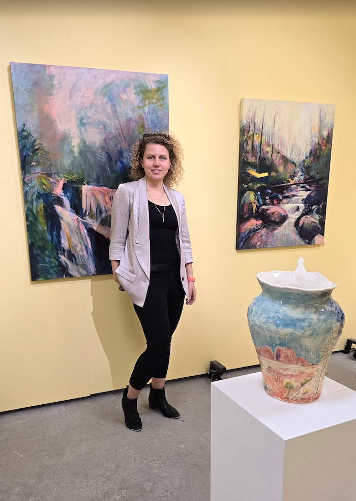 A woman standing in front of two paintings of nature scenes in an art gallery, with a large ceramic vase on a white pedestal nearby.