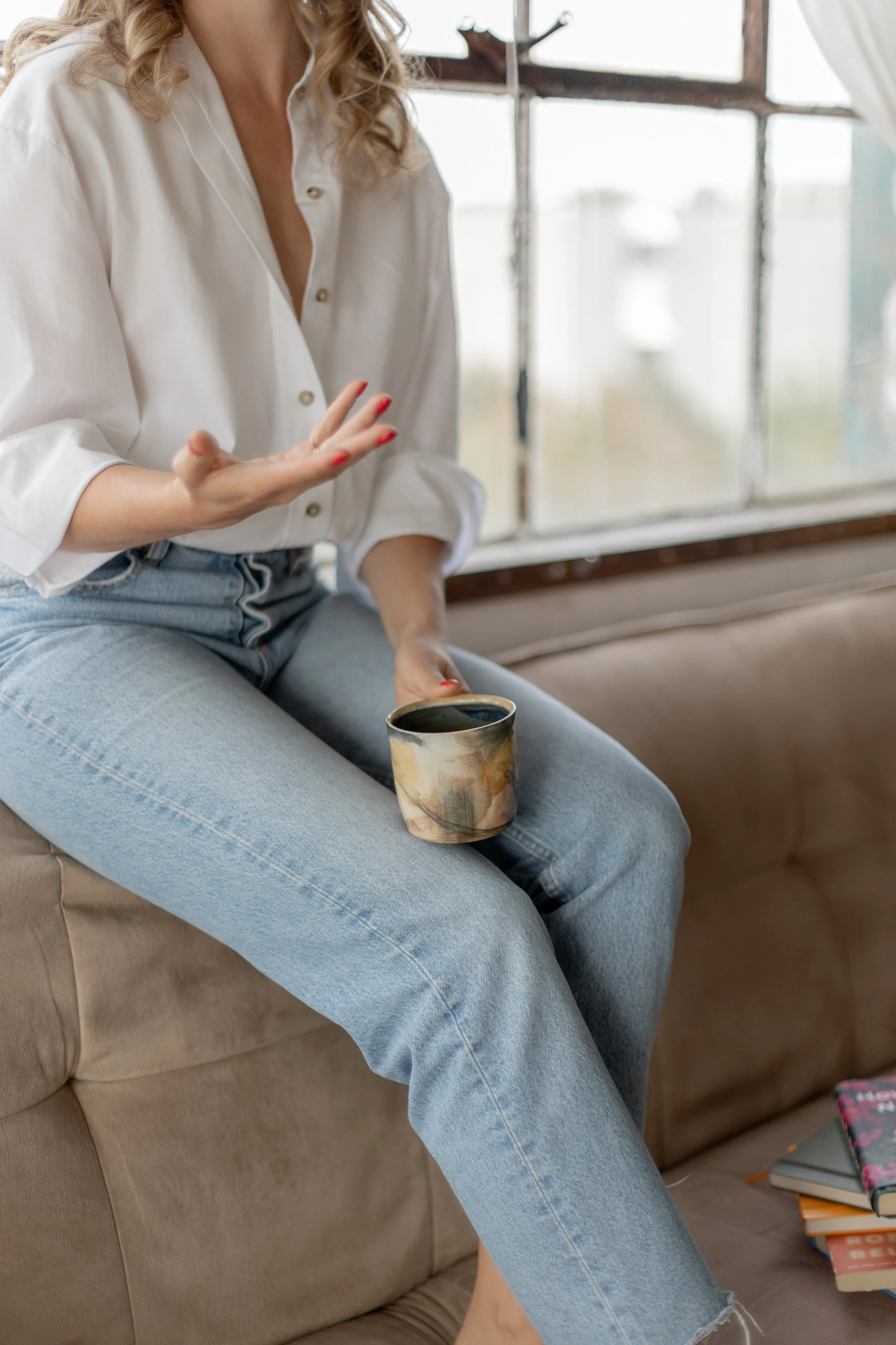 A close up of Kate sitting on the back of the couch, with a ceramic mug on her lap in one hand and her other hand in motion while she talks