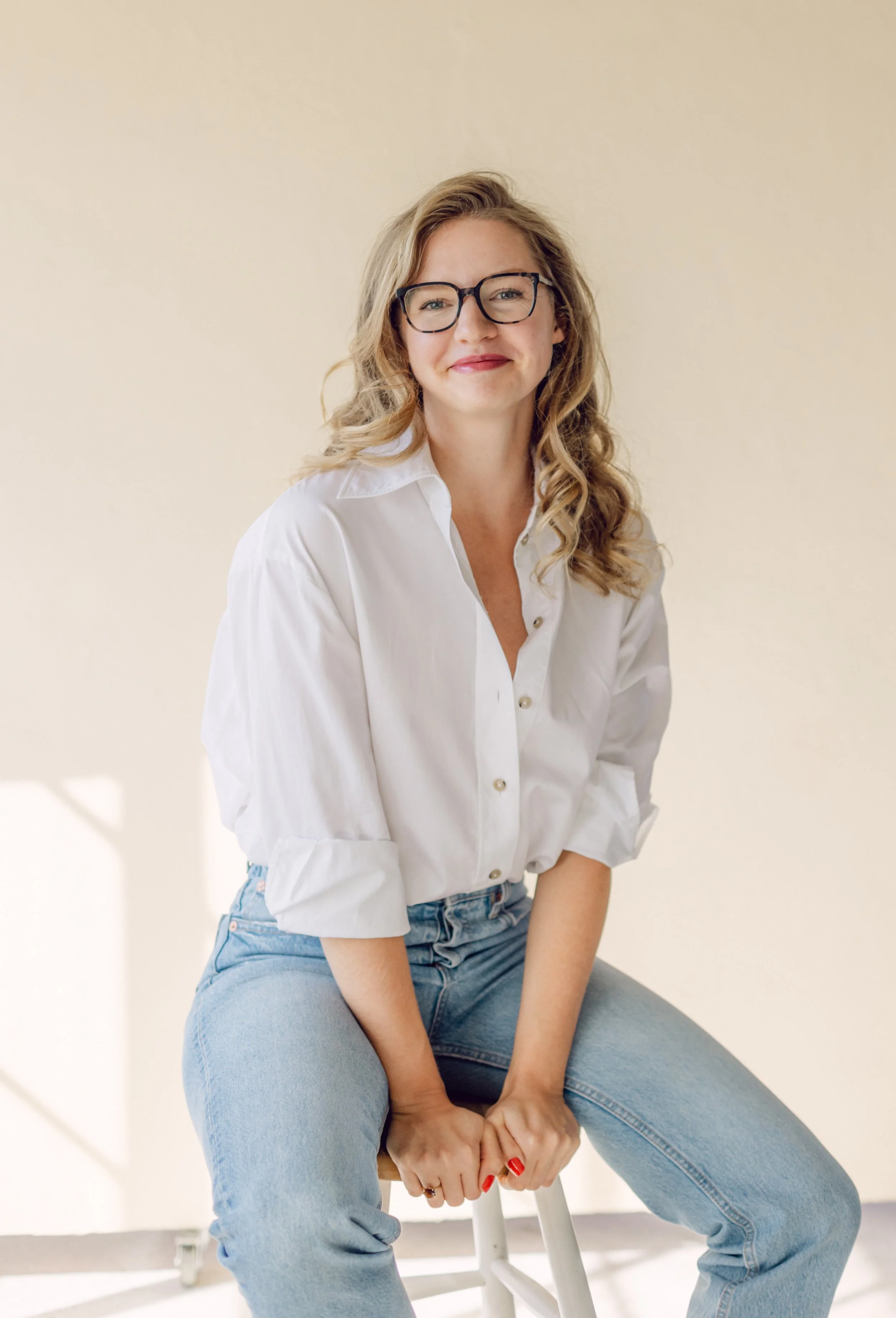 Kate, a white woman with dark glasses and curly dark blonde hair, sits on a stool in an art studio and smiles directly into the camera