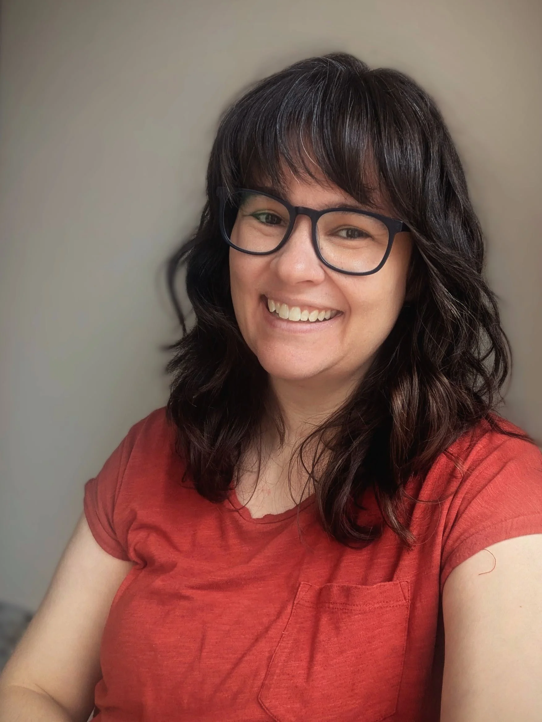 A white woman with dark, wavy hair and bangs smiles for the camera. She wears glasses and a red tee.