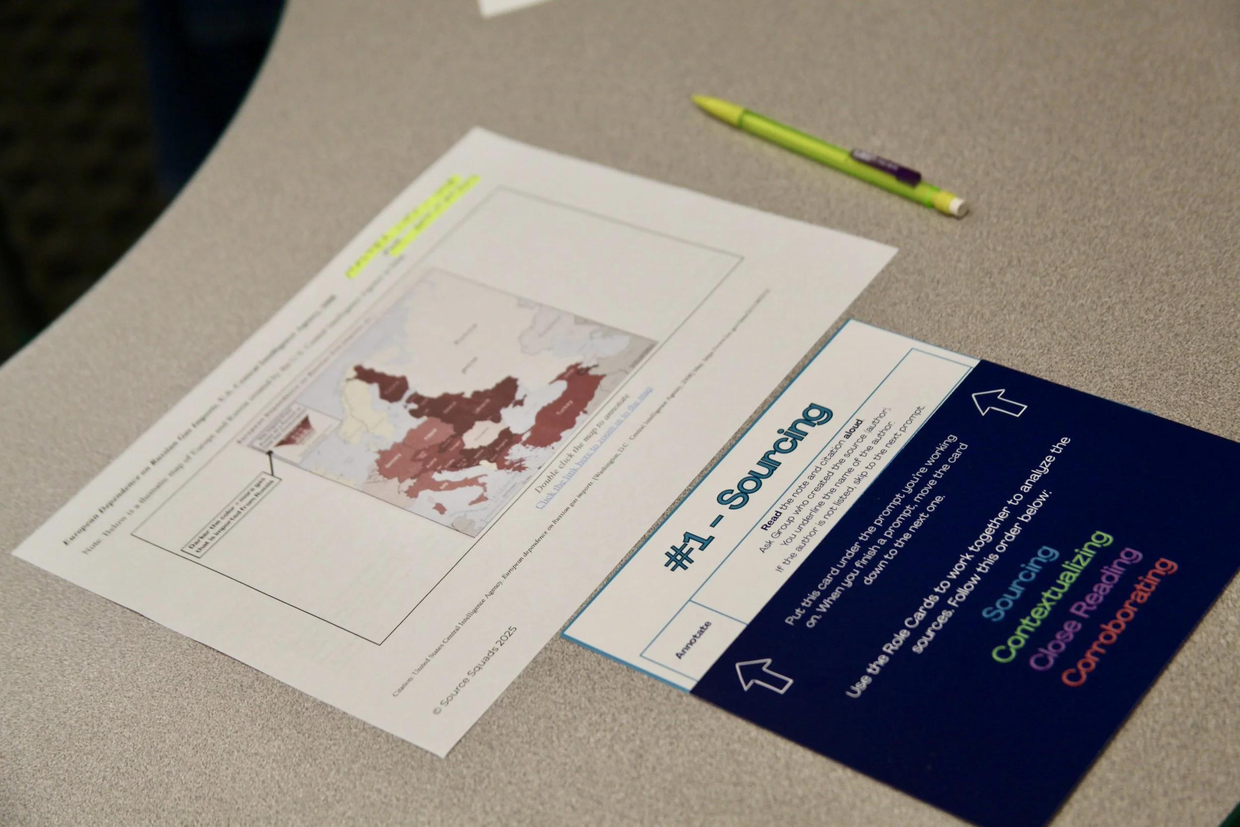 A gray table with a printed sheet of paper showing a color-coded map of Europe indicating various countries in different shades of red and beige, a green pen, and a blue informational flyer with instructions.