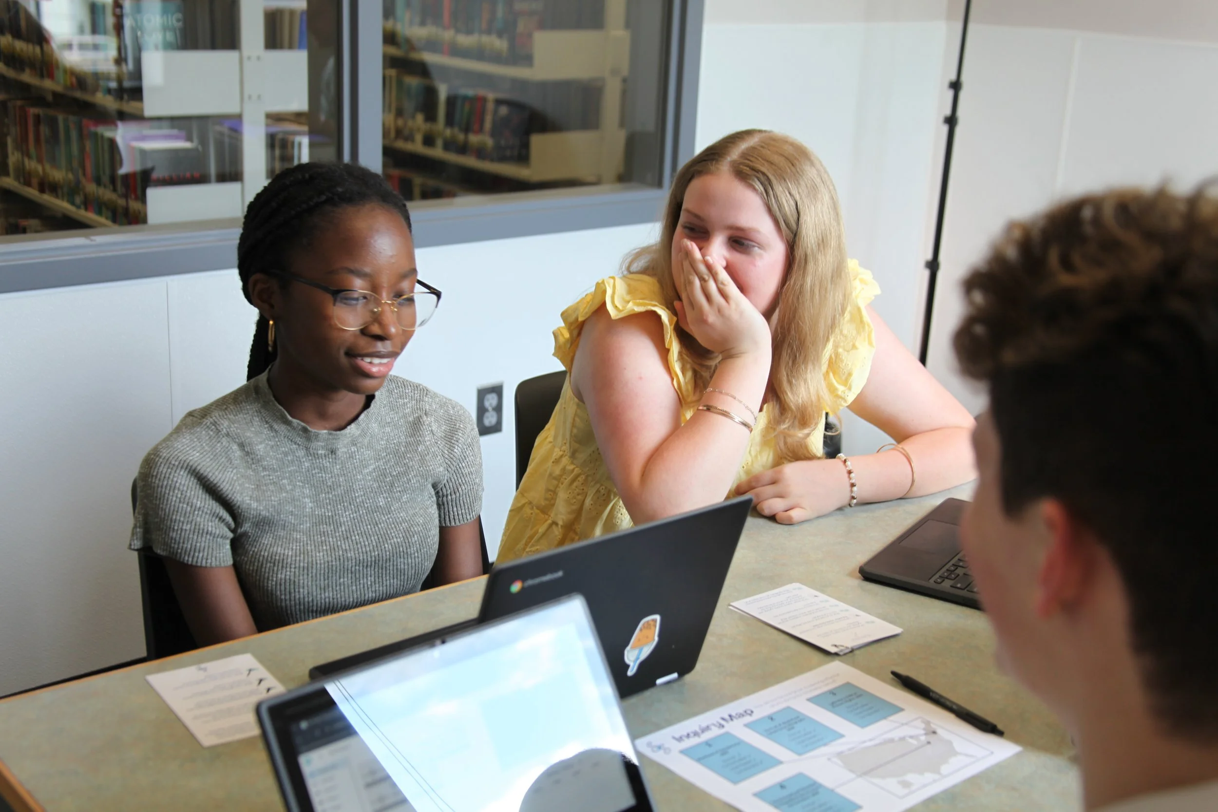Four students sit around a table in a study room, engaged in conversation. They have laptops, papers, and a pen on the table. One girl in a yellow dress appears surprised or amused, while another girl with glasses looks at her laptop. Two other students are partially visible, focusing on the discussion.