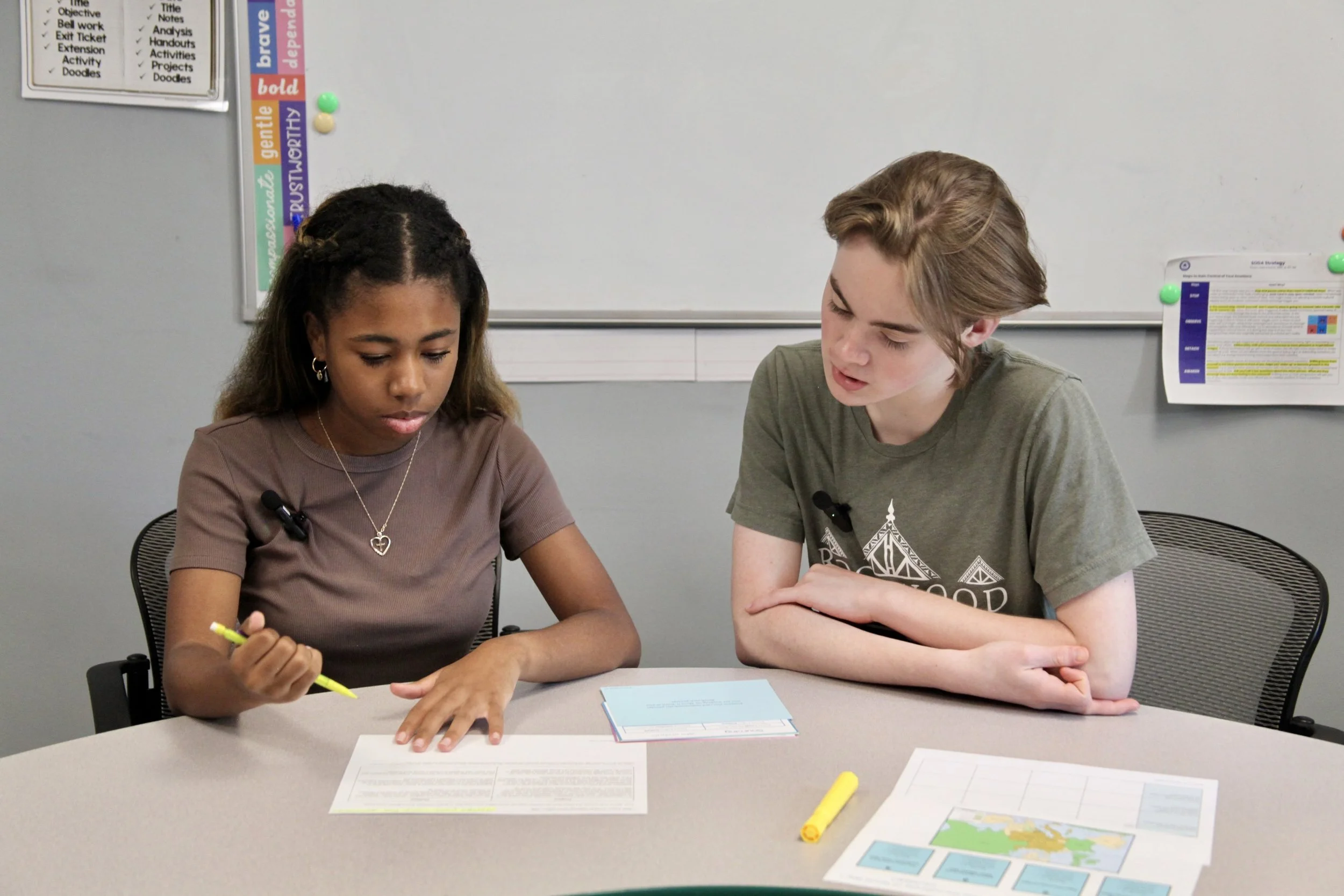 Two young students, a girl with dark hair and a boy with light brown hair, sit at a table working together on a project or worksheet. They are focused, with papers, a map, and highlighters on the table in front of them. They are in a classroom setting, with a whiteboard and educational posters on the wall behind them.