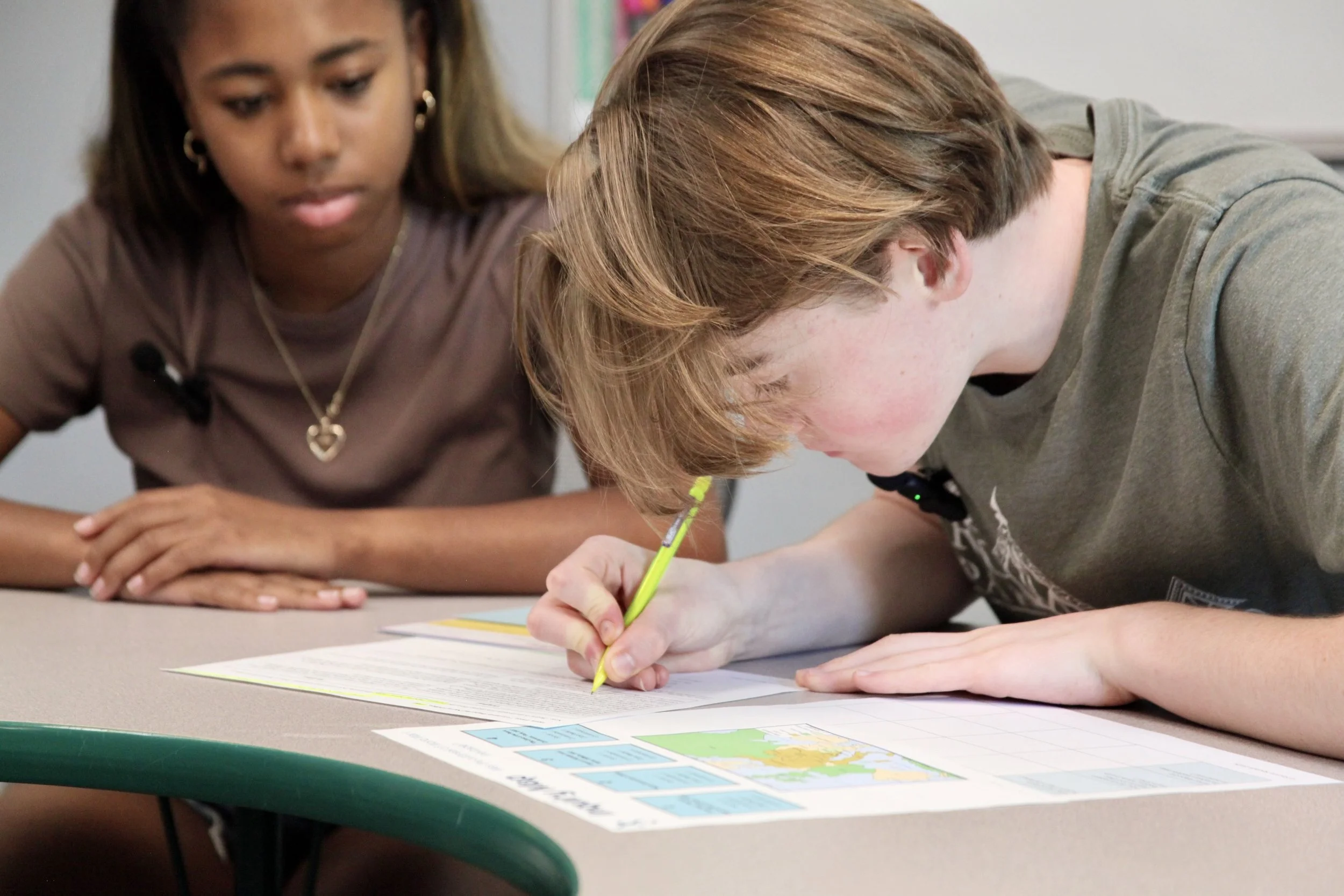 A teenager with brown hair is writing on a piece of paper with a yellow pencil while a woman with darker skin and long earrings watches. They appear to be working on a school assignment or activity.