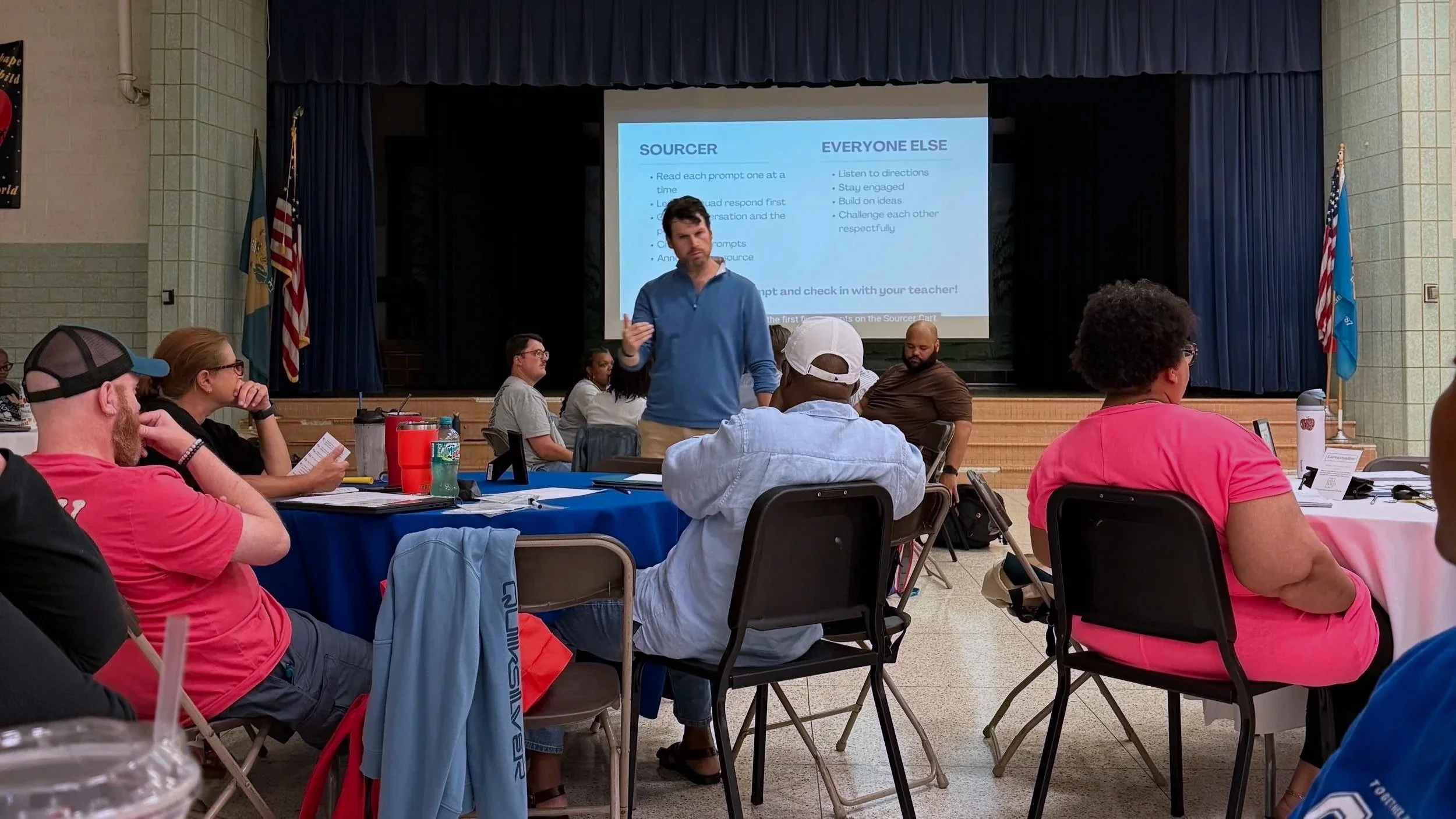 A man standing in front of a projector screen giving a presentation to a seated audience in a large room. The screen displays a PowerPoint slide with instructions for a classroom activity.