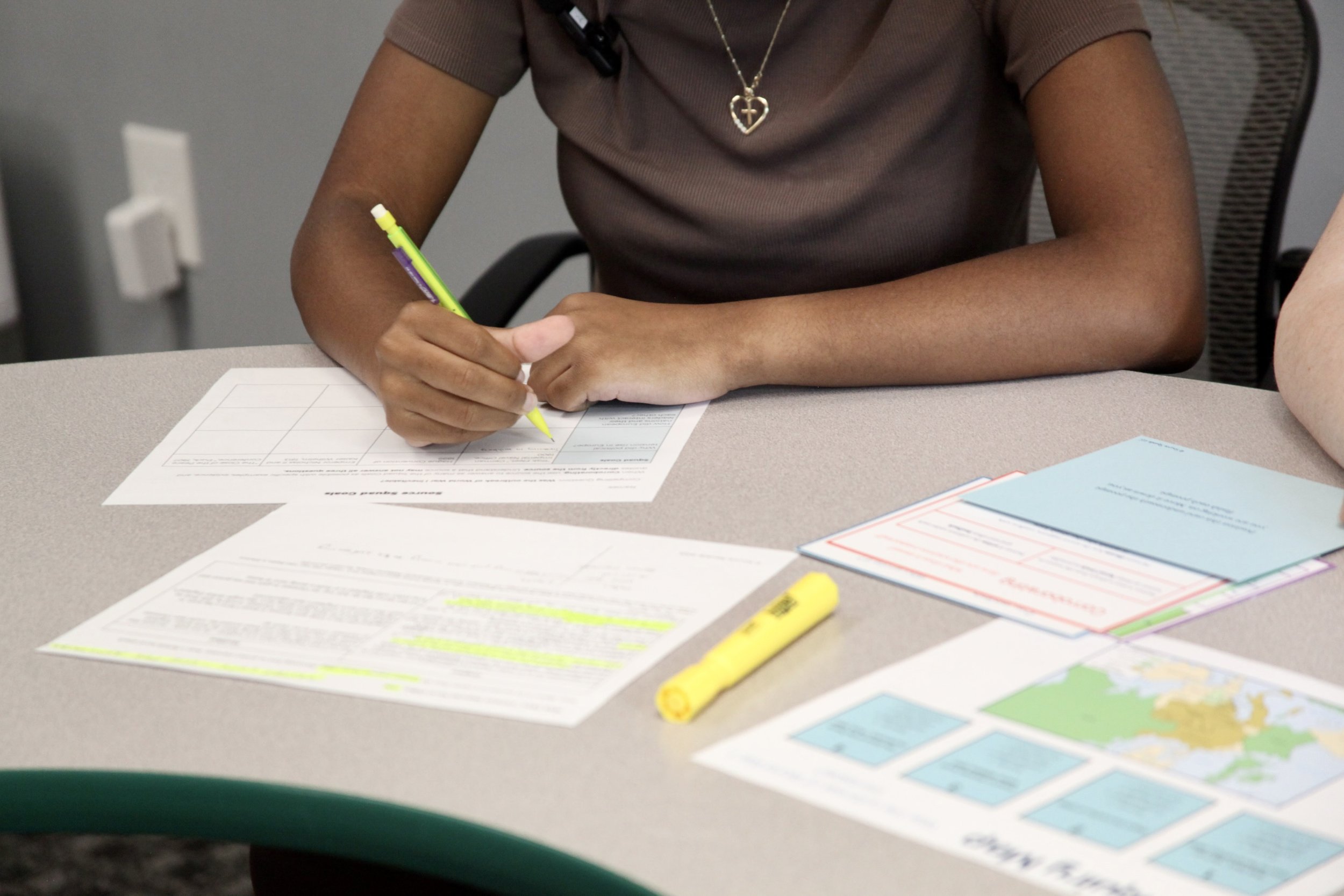 Person wearing a brown shirt is seated at a table, filling out paperwork with a yellow highlighter and a green pen. There are color-coded maps or charts and highlighters on the table.