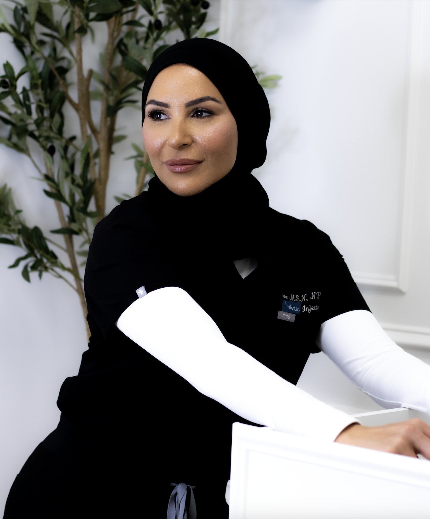 Vrouw in medische outfit met hoofddoek, zittend achter een balie, binnen een kliniek of kantoorruimte, met groene planten op de achtergrond.