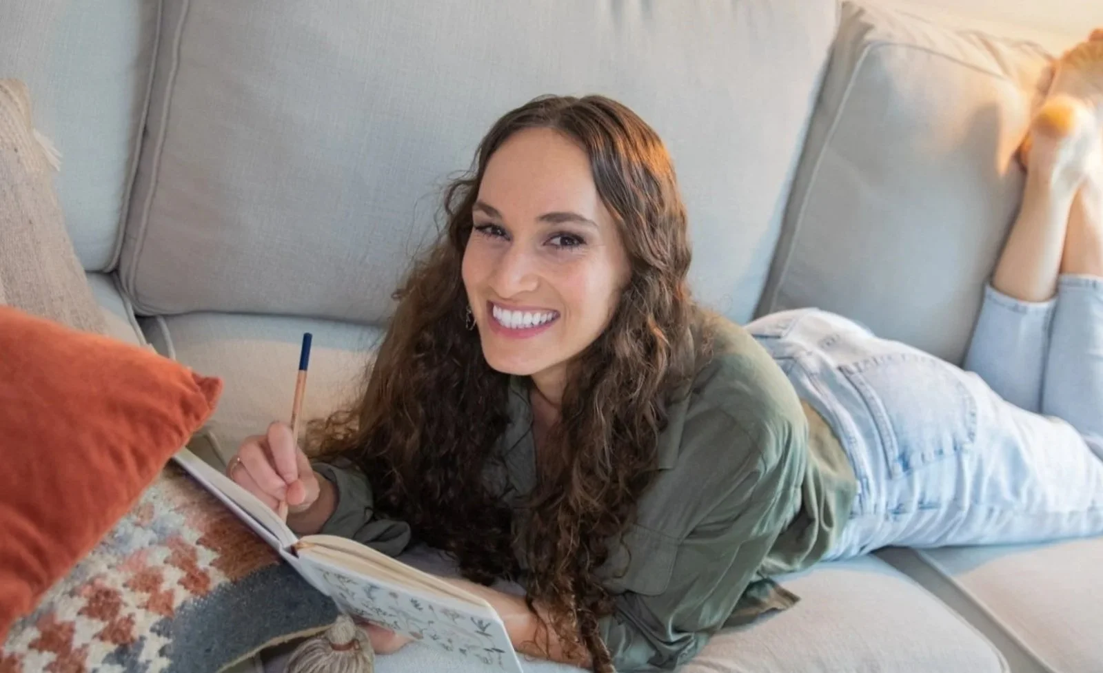 Photo of Maggie Goulder -- web designer for therapists -- laying on her stomach on a couch, smiling and holding a pencil and a notebook.