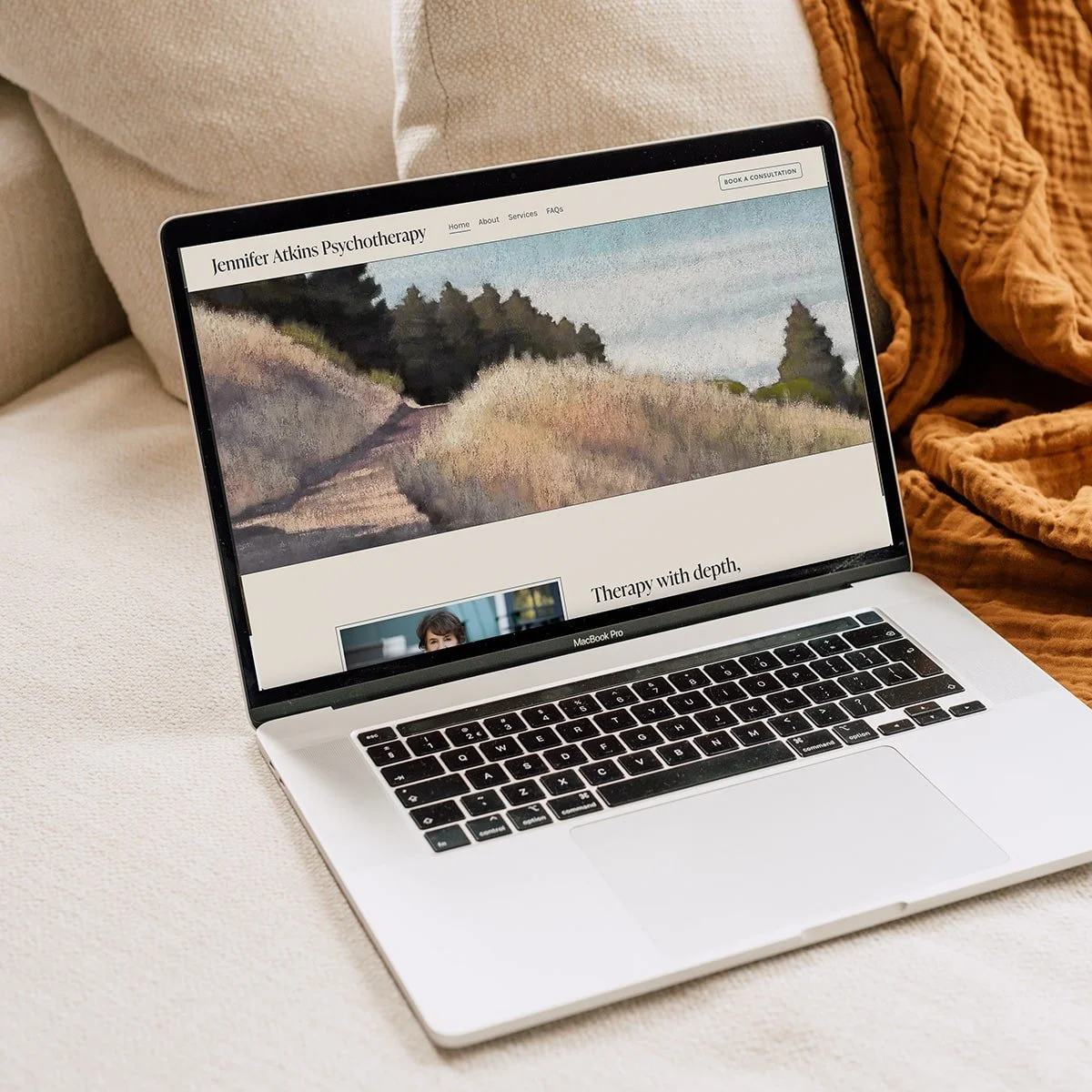 A MacBook Pro laptop on a beige couch displaying a psychotherapy website with scenic artwork and a photo of a woman.