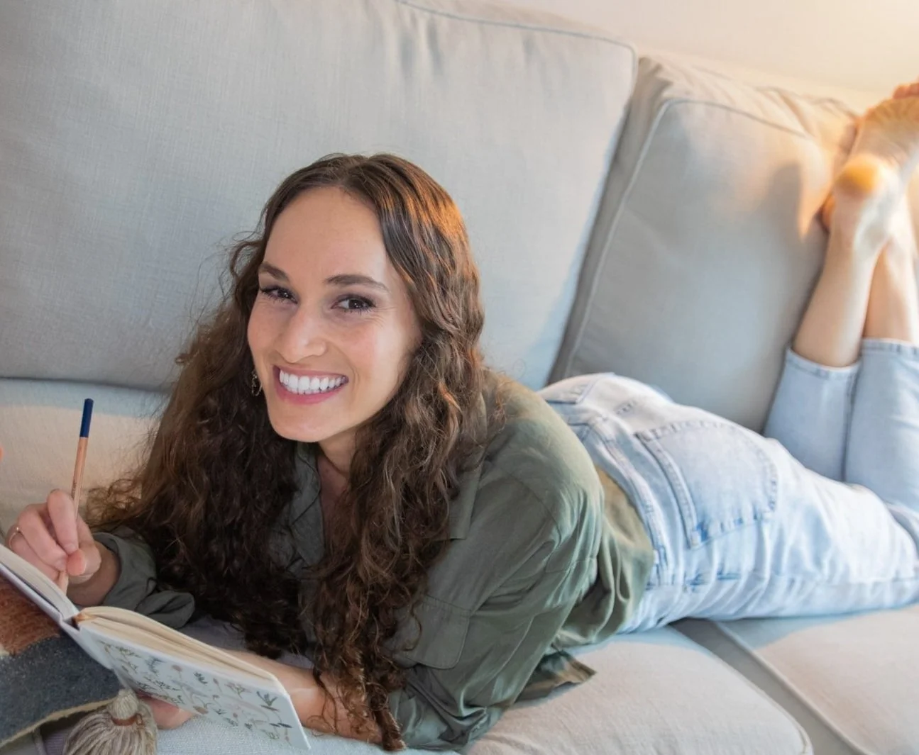 Maggie Goulder, custom web designer for therapists, lying on her stomach on a couch, smiling, holding a pen, and writing in a notebook.