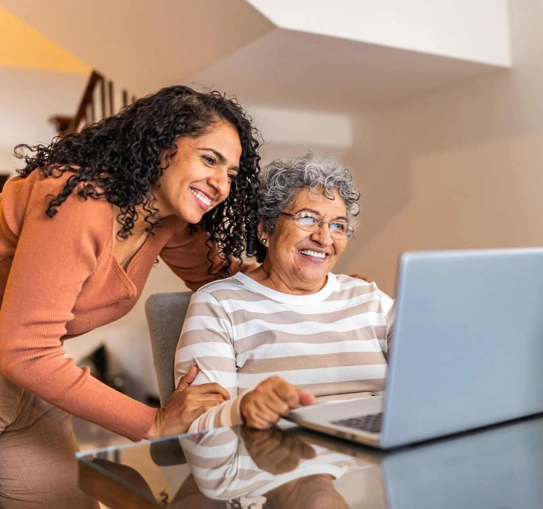 Two women, one younger and one older, smiling and looking at a laptop screen together in a cozy home setting.
