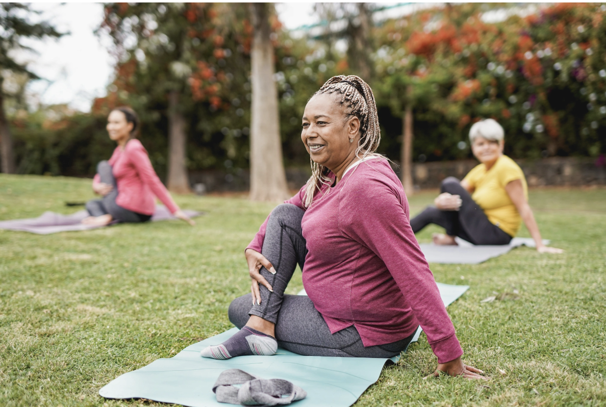 Three women of different ages and backgrounds smiling and stretching on yoga mats during an outdoor yoga session in a garden setting, representing the accessible, inclusive, and community-centered yoga and somatic practices offered through Malachi Gi