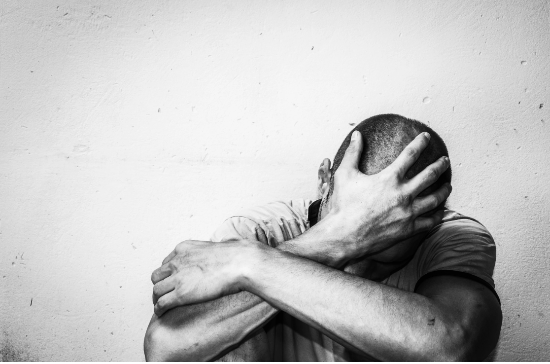 A black and white photograph of a man sitting against a wall with his head bowed into his hands in a posture of deep emotional distress, representing the pain and isolation that many trauma survivors experience before seeking support through Malachi
