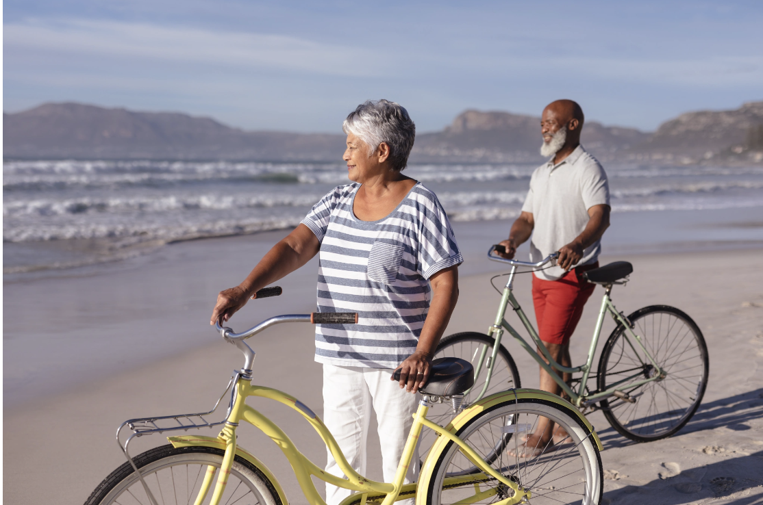 An older couple walking their bicycles along a sunny beach with a relaxed and contented demeanor, representing the renewed sense of joy, connection, and relational wellbeing that can emerge through the trauma and family counseling work offered by Mal