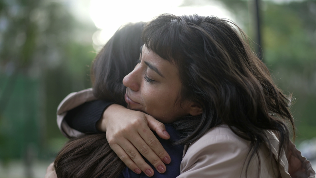 Two women sharing a close, heartfelt embrace outdoors with soft green bokeh in the background, conveying the emotional safety, human connection, and relational healing at the core of Malachi Gillihan's trauma recovery work