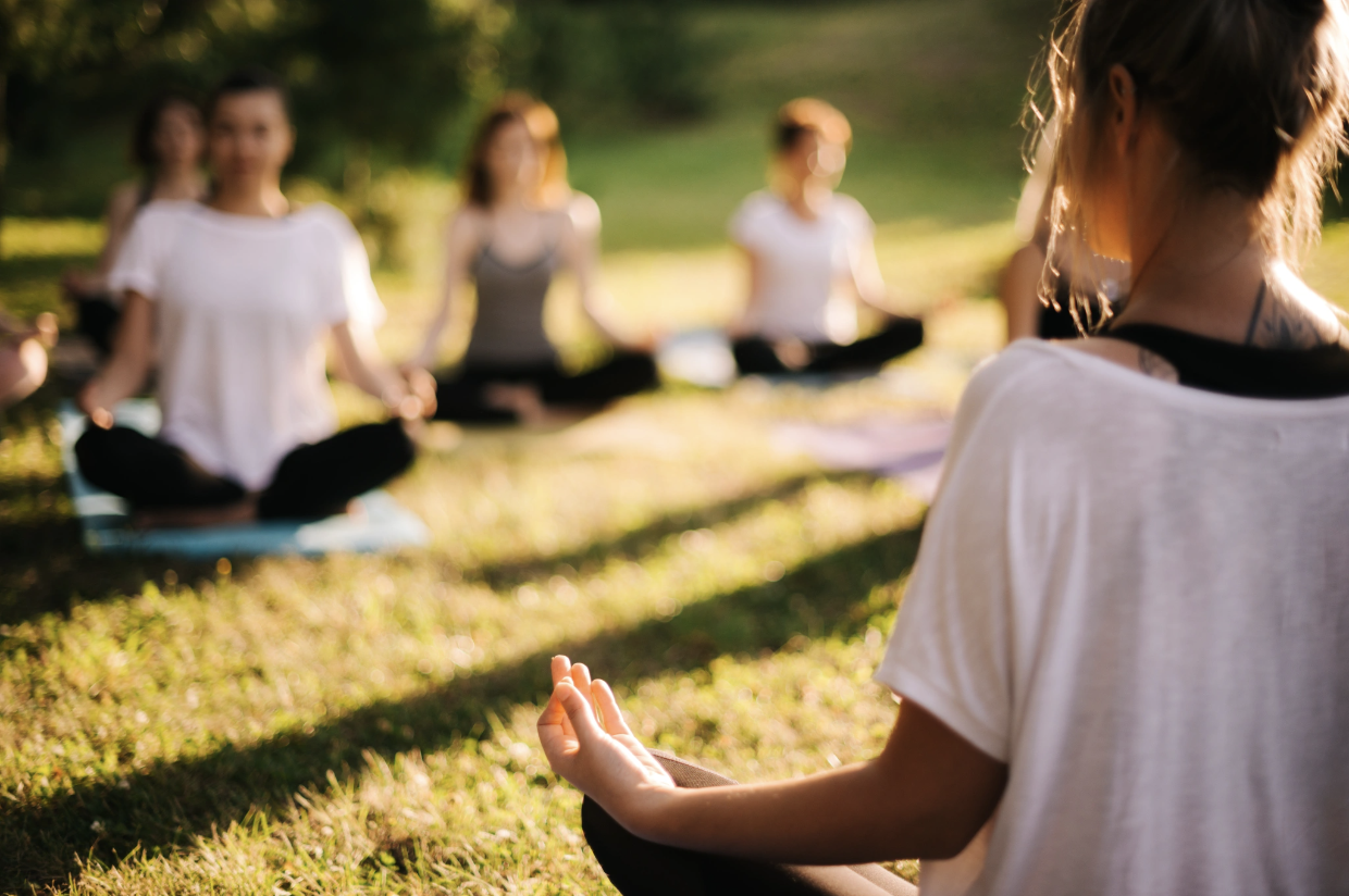 A group of people seated in a meditative yoga pose on grass in soft natural light, representing the somatic and mind-body practices Malachi Gillihan integrates into his trauma recovery work