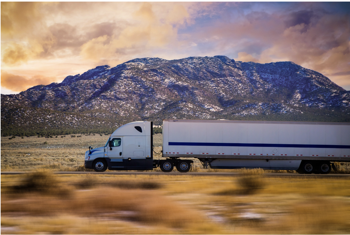 semi-truck traveling along a desert highway with a dramatic mountain range and stormy sunset sky in the background