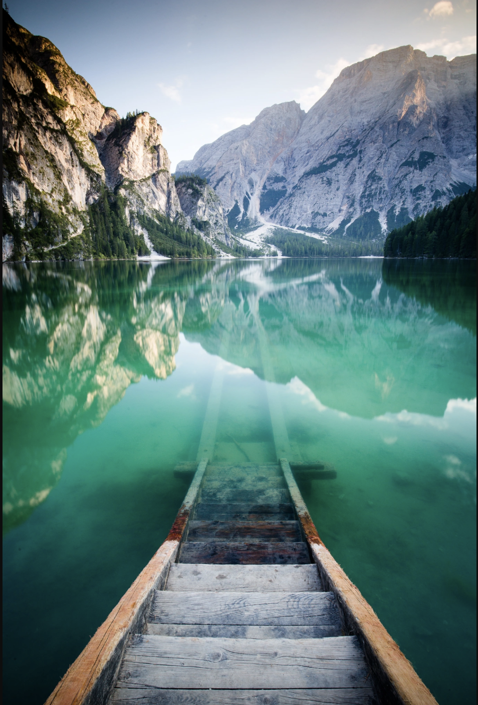 Weathered wooden steps leading down into a still turquoise alpine lake with dramatic mountain peaks reflected in the water, evoking the courage it takes to step into the healing process and the clarity and depth that await within Malachi Gillihan's