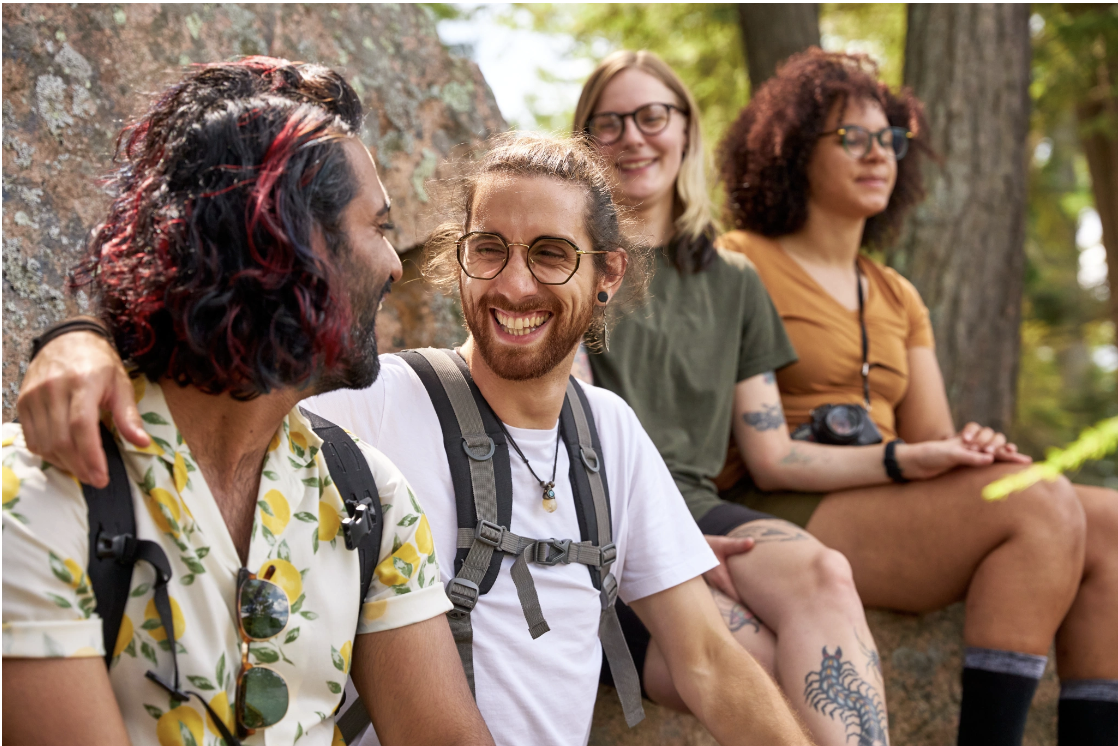 A diverse group of young adults laughing and relaxing together outdoors in a forest setting with backpacks, conveying the joy, authentic connection, and sense of belonging that Malachi Gillihan fosters through his group trauma recovery programs