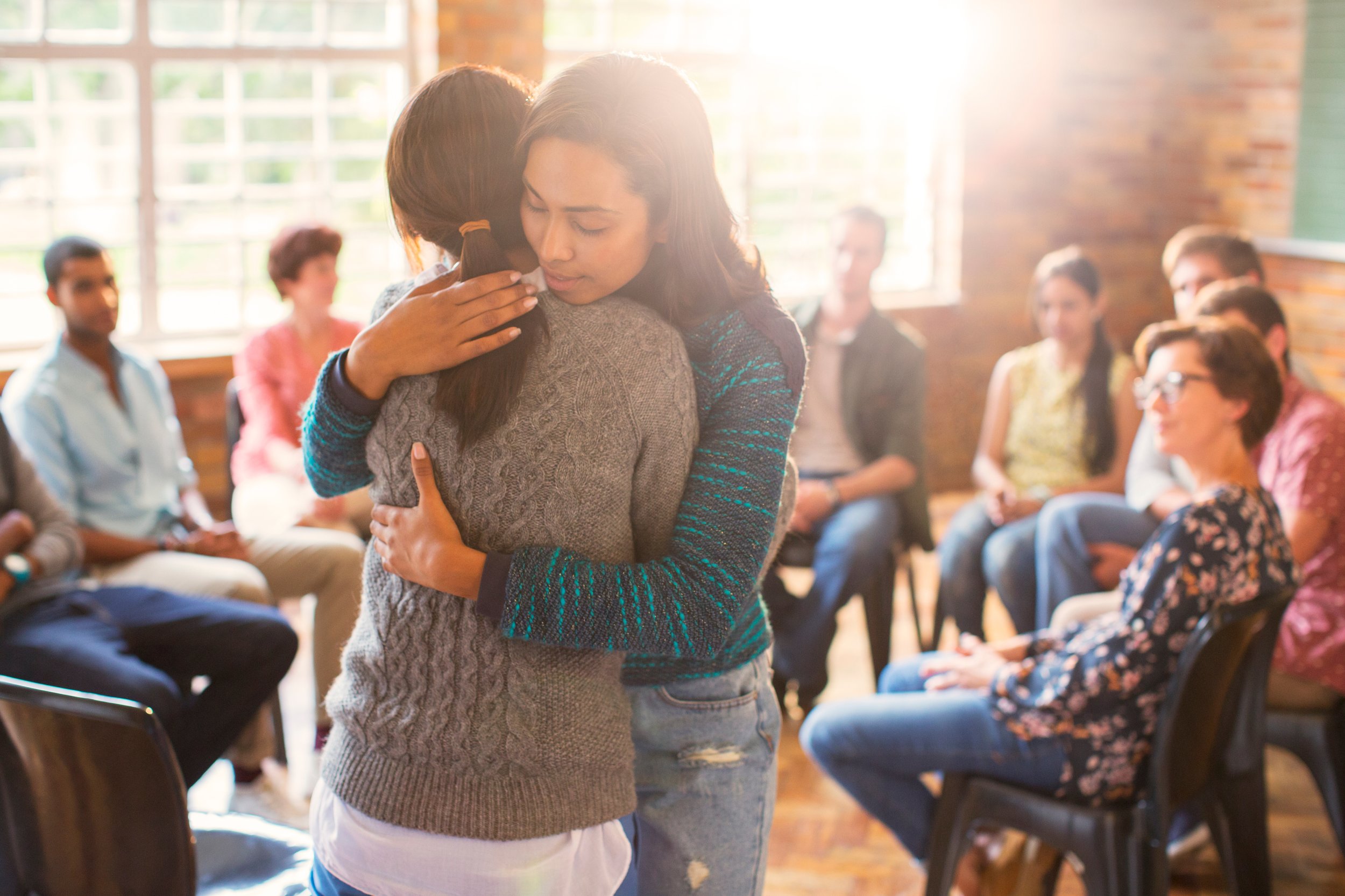Two participants sharing a supportive embrace during a group therapy session, with other members seated in a circle in the background, reflecting the safety and human connection at the heart of Malachi Gillihan's group trauma recovery programs