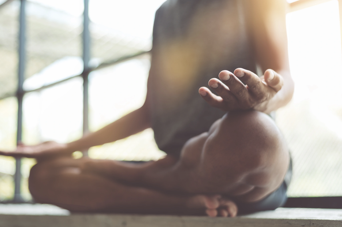 Close-up of a man seated cross-legged in a meditative pose with hands resting on his knees in warm natural light, representing the mindfulness and meditation practices Malachi Gillihan integrates into his trauma recovery work