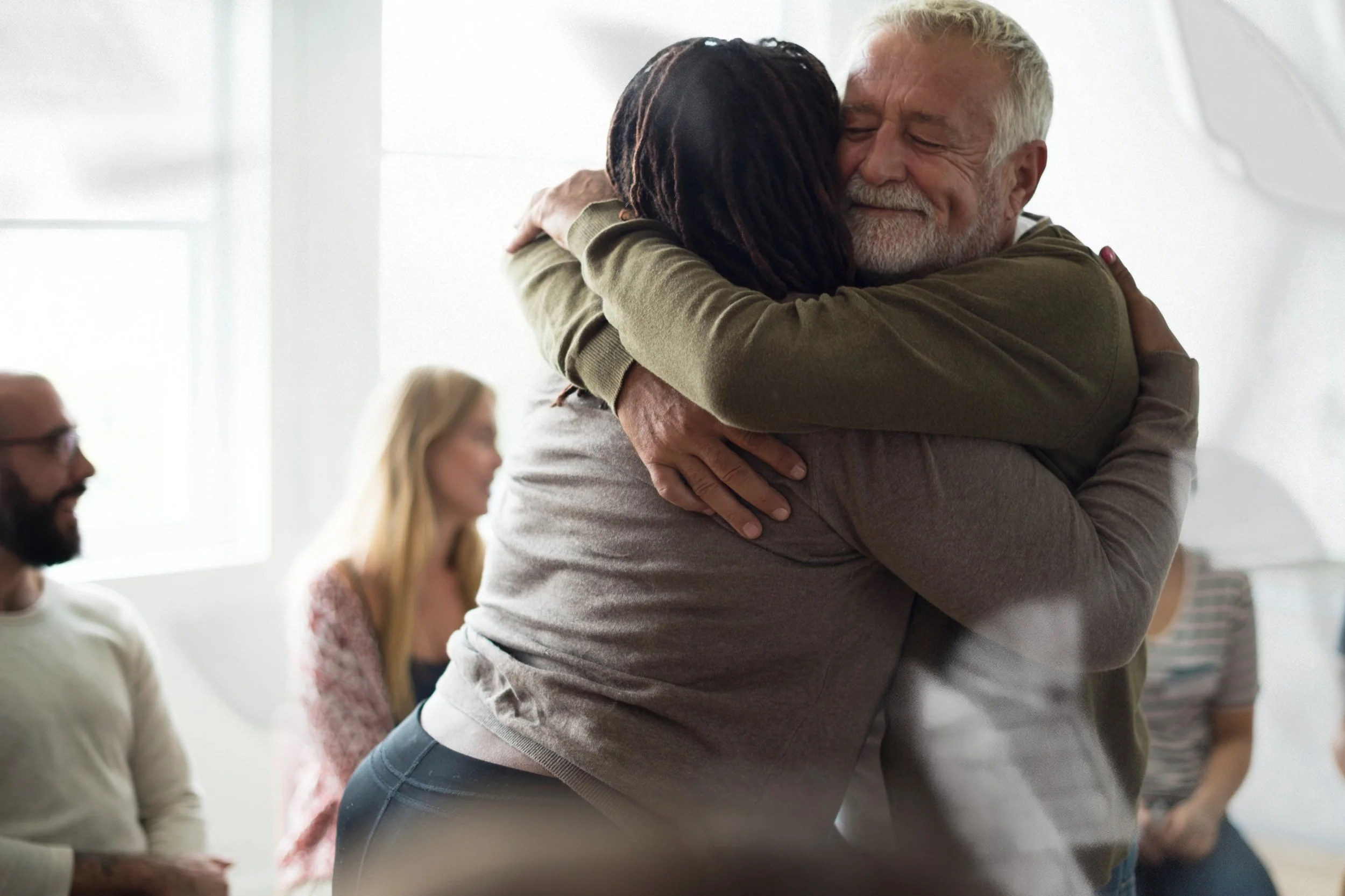 Two people sharing a warm embrace during a group therapy session while others look on with smiles, capturing the human connection, trust, and emotional safety that Malachi Gillihan cultivates in his trauma recovery group programs