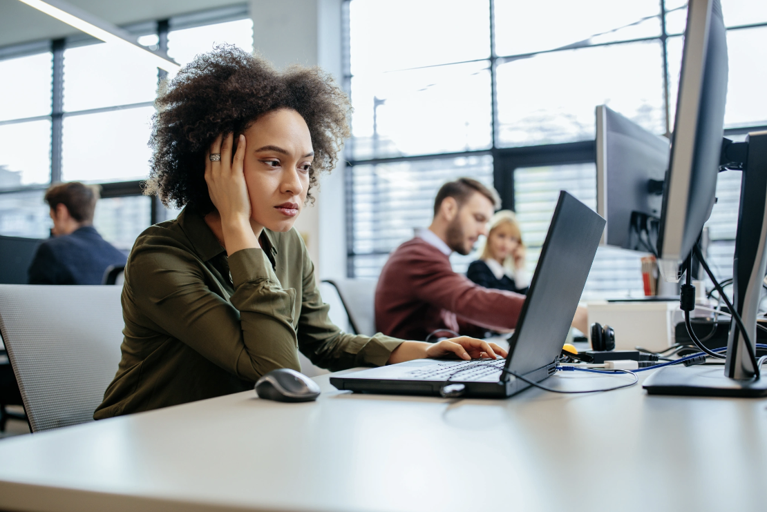 A woman sitting at a workplace desk looking fatigued and distracted while working on a laptop, illustrating how unresolved trauma can affect concentration, energy, and career performance