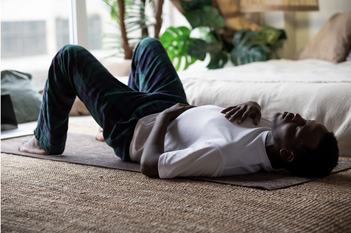 A man lying on a yoga mat with knees bent and hands resting on his chest in a grounded breathwork position, representing the somatic and nervous system regulation practices that Malachi Gillihan integrates into his trauma recovery work