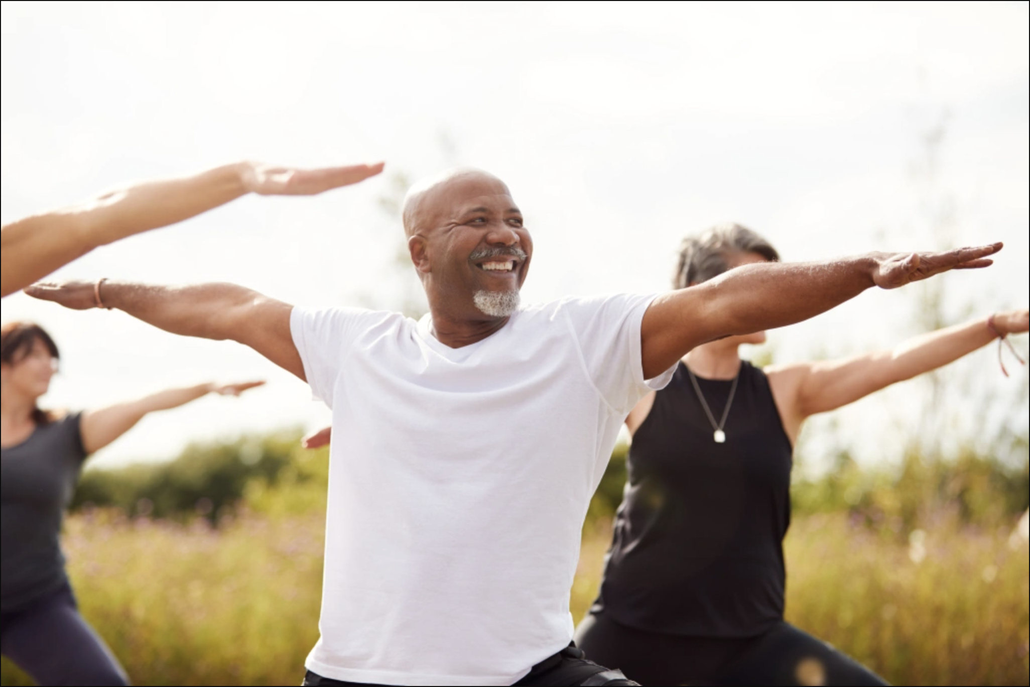 An older man smiling broadly with arms extended in warrior pose during an outdoor group yoga class in a sunlit meadow, representing the joy, embodiment, and inclusive mind-body healing practices central to Malachi Gillihan's trauma recovery work