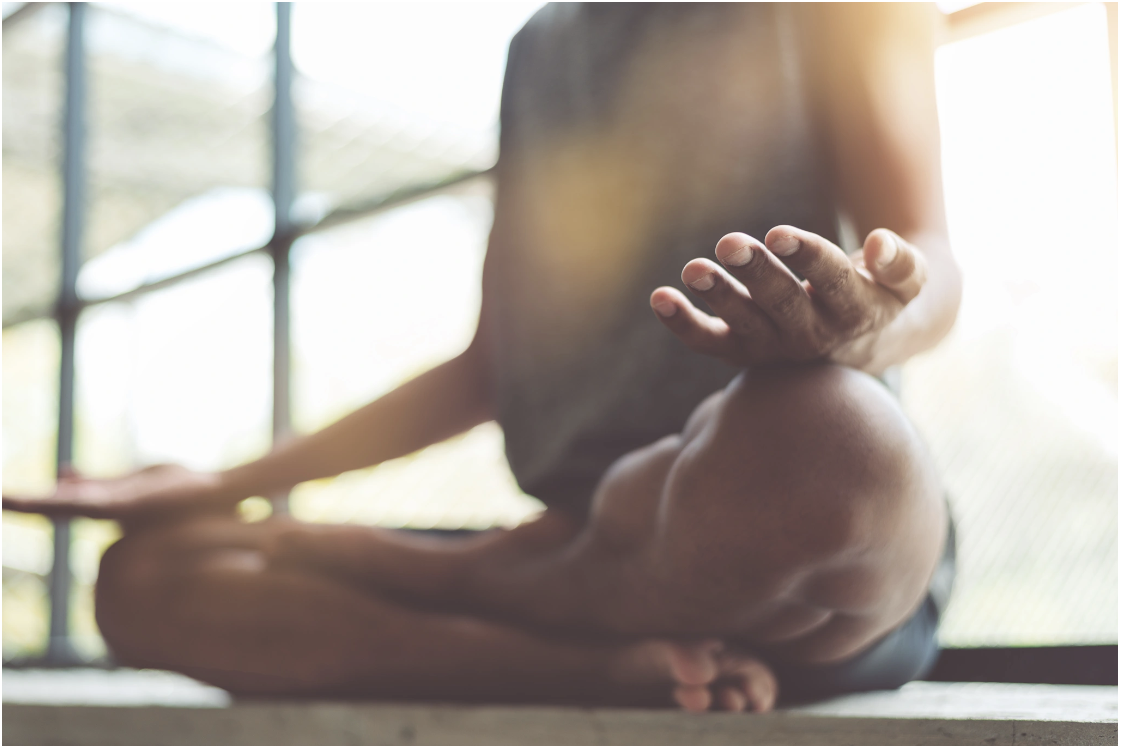Close-up of a man seated in a cross-legged meditation pose with hands resting on his knees in warm backlit natural light, representing the mindfulness and meditation practices Malachi Gillihan integrates into his trauma recovery work