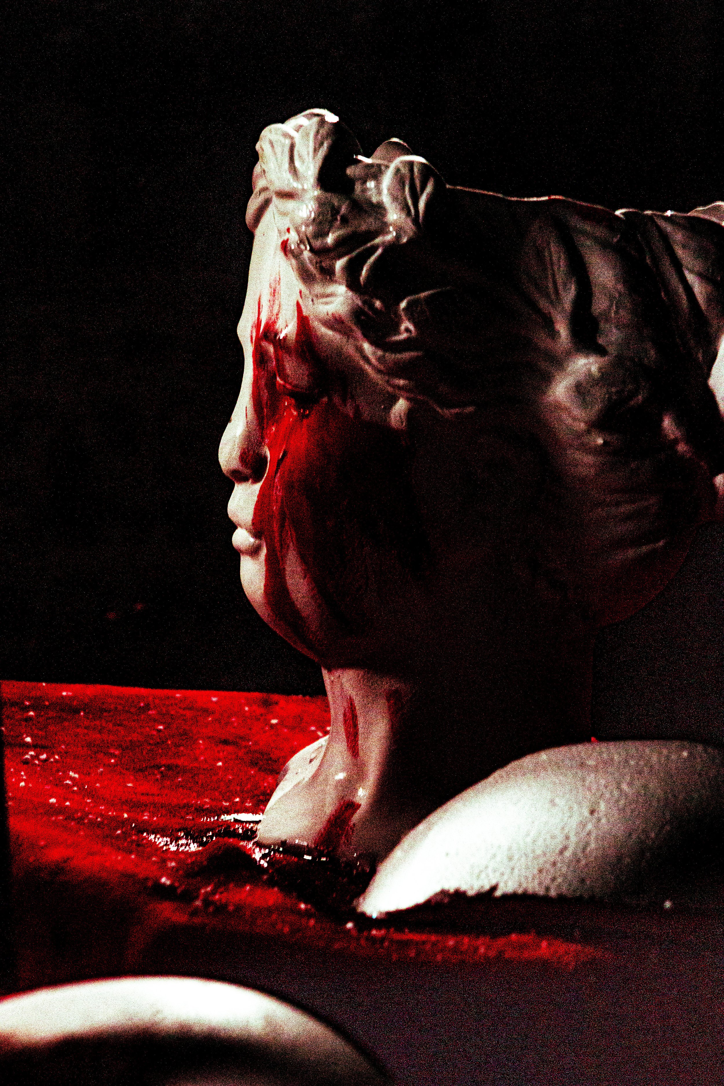Bust statue of a woman with red paint splattered on the face against a dark background.